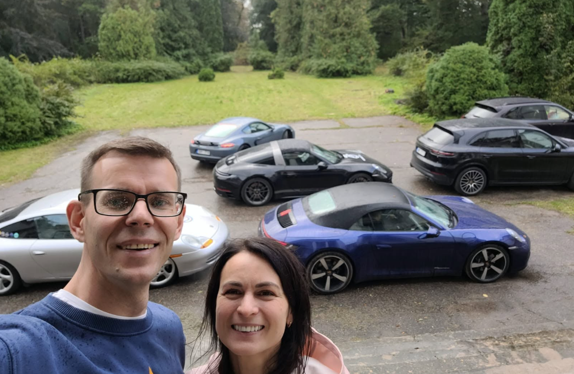 Two smiling people taking a selfie in front of multiple parked cars, with a grassy area and trees in the background.