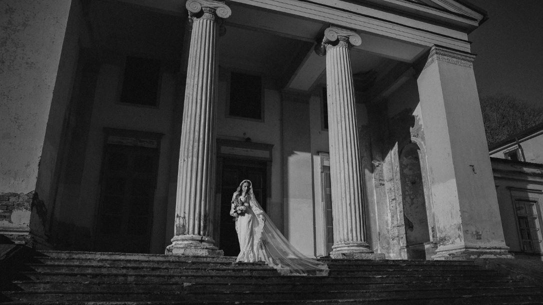 Black and white photo of a woman in a wedding dress holding a bouquet, standing on the stairs in front of an old building with tall columns.