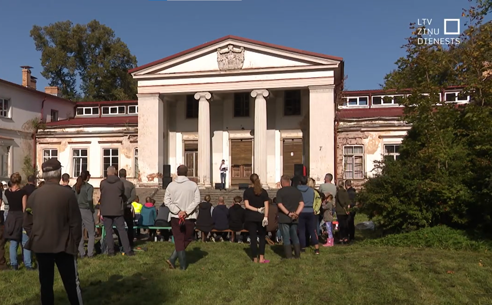 A crowd gathered in front of an old, weathered building with columns, some people are standing while others are seated on benches, and a person is speaking or performing on a small stage in front of the building.