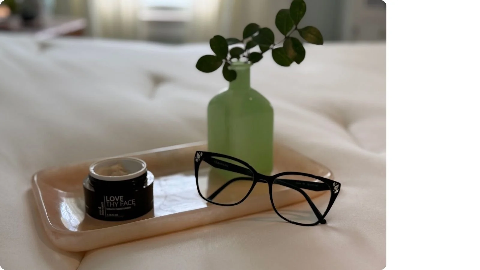 Open jar of Love Thy Face Miracle Moisturizer beside black reading glasses and a pale green glass vase holding a fresh crepe myrtle sprig on a blush marble tray atop a white quilted bed.