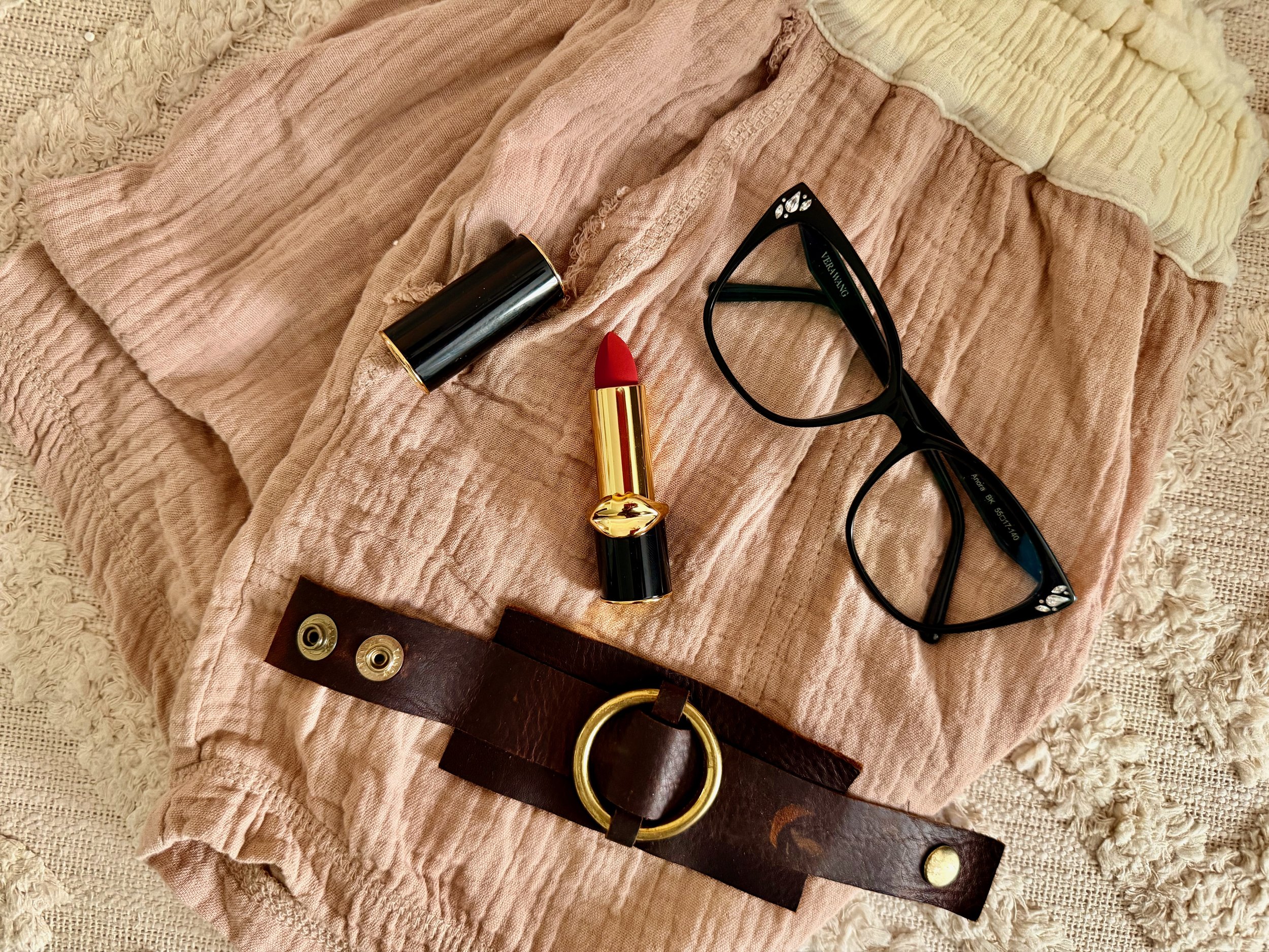 A worn leather bracelet, lipstick, black glasses, and soft textured fabric arranged as a lived-in style flat lay.