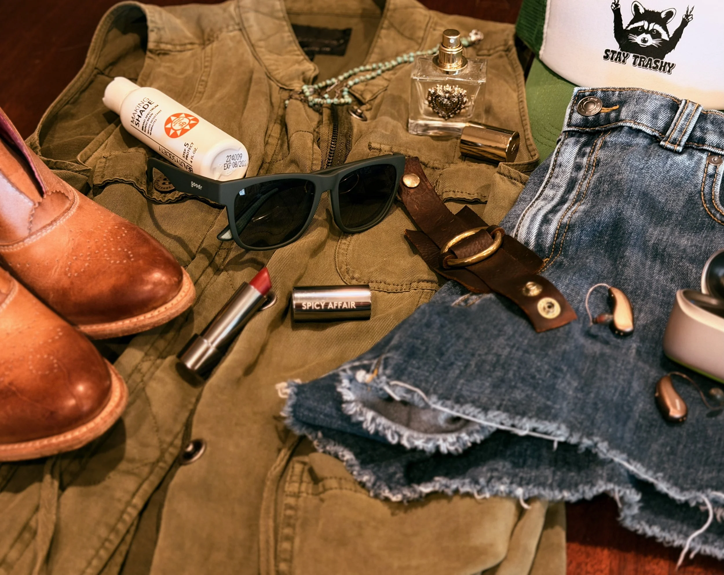 A flat lay of hearing aids, lipstick, sunglasses, denim, leather boots, and worn accessories representing real-life personal style.