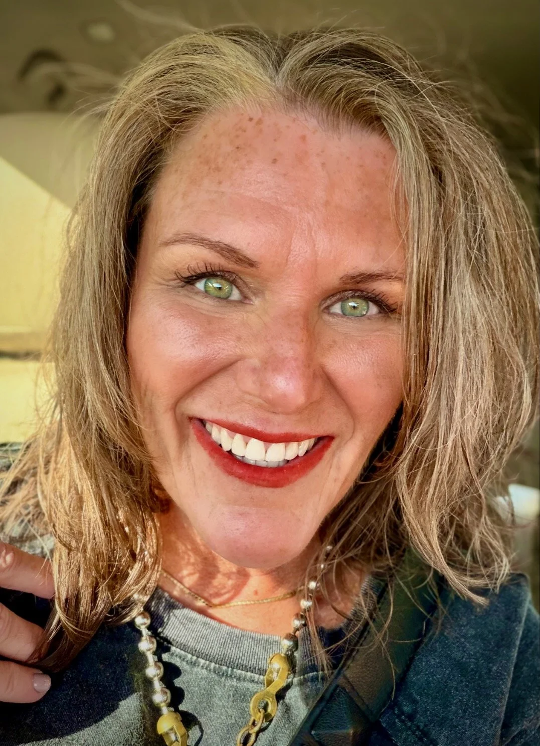 Julie smiling in natural light with bare skin, lipstick, and everyday jewelry.