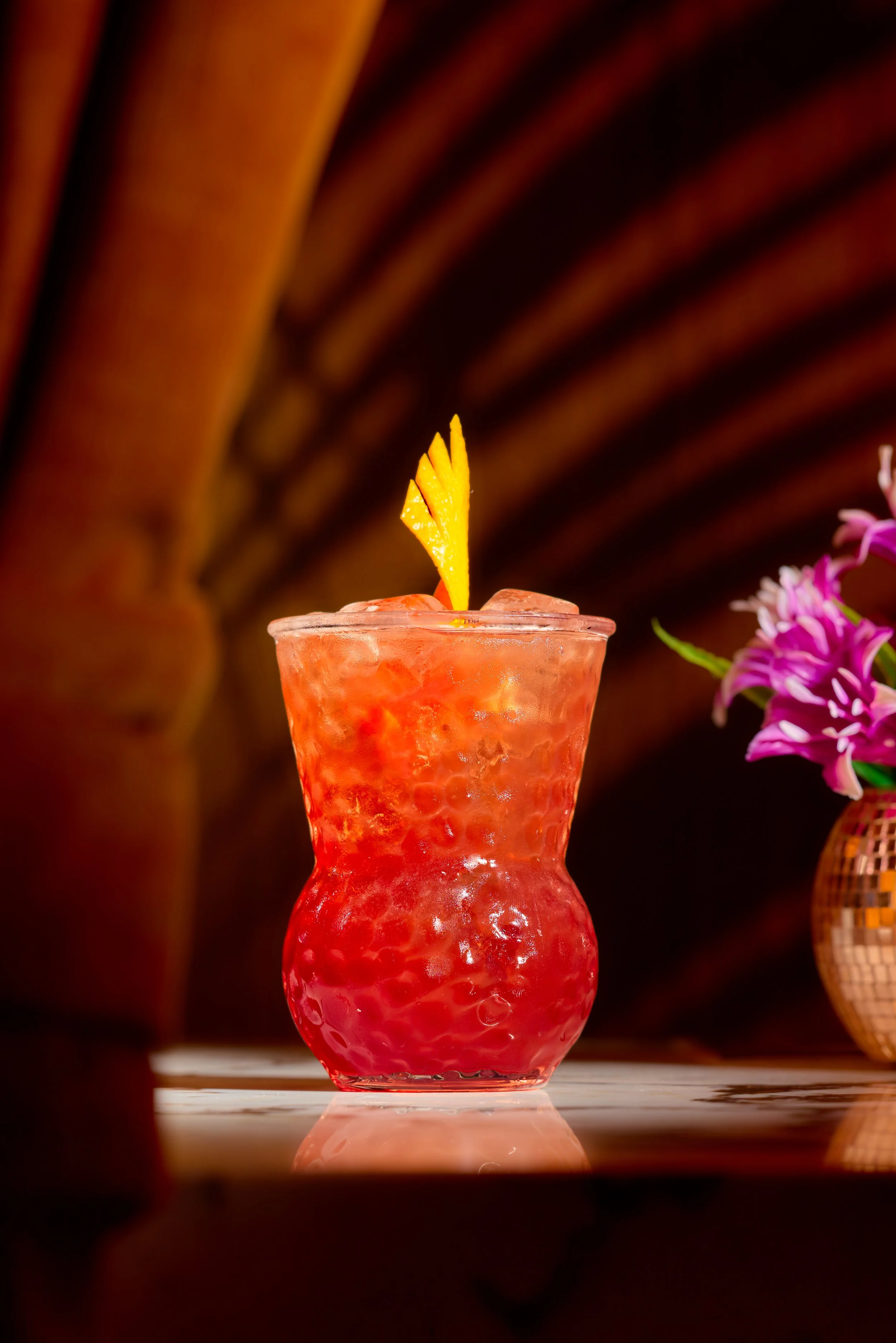 A pinkish-red cocktail with ice in a decorative glass, garnished with a yellow fruit slice and a small yellow decorative element, set against a dark background with a purple flower on the right.