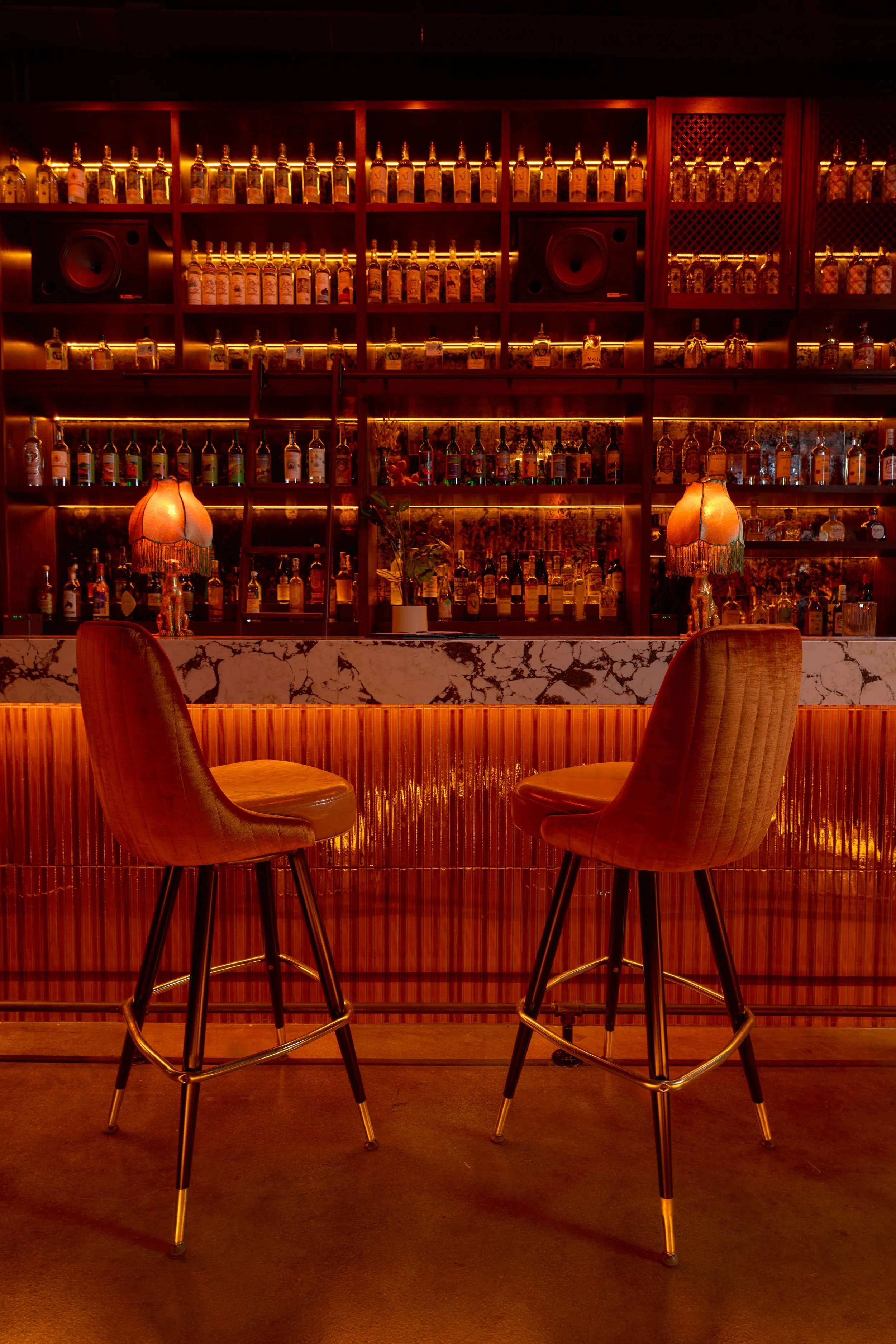 Interior of a bar with a wooden counter, two high chairs, and shelves filled with bottles of alcohol illuminated with warm lighting. Two vintage-style lamps with fringed shades are on the bar counter.