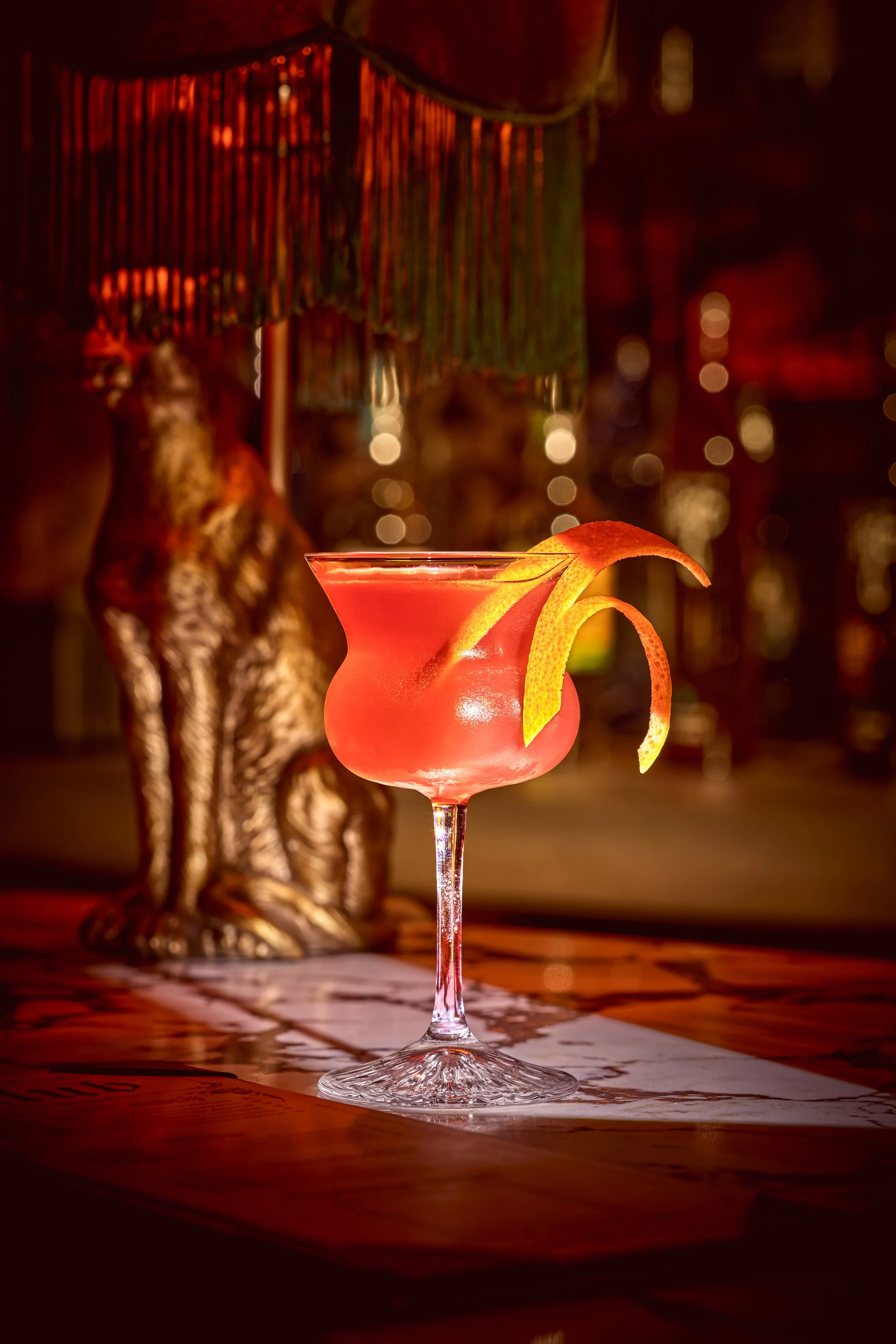 A pink cocktail in a stemmed glass garnished with a lemon twist, in a dimly lit setting with a tasseled lamp and a decorative statue of a lion in the background.