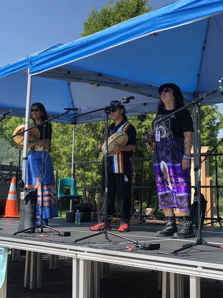 Three women on a stage singing into microphones, dressed in colorful clothing, with greenery and trees in the background.