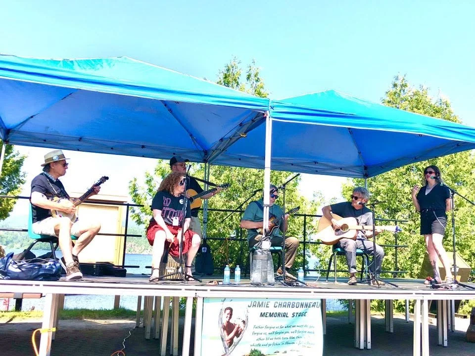 A group of six musicians performing on a stage outdoors under a blue canopy, with trees and a clear sky in the background.