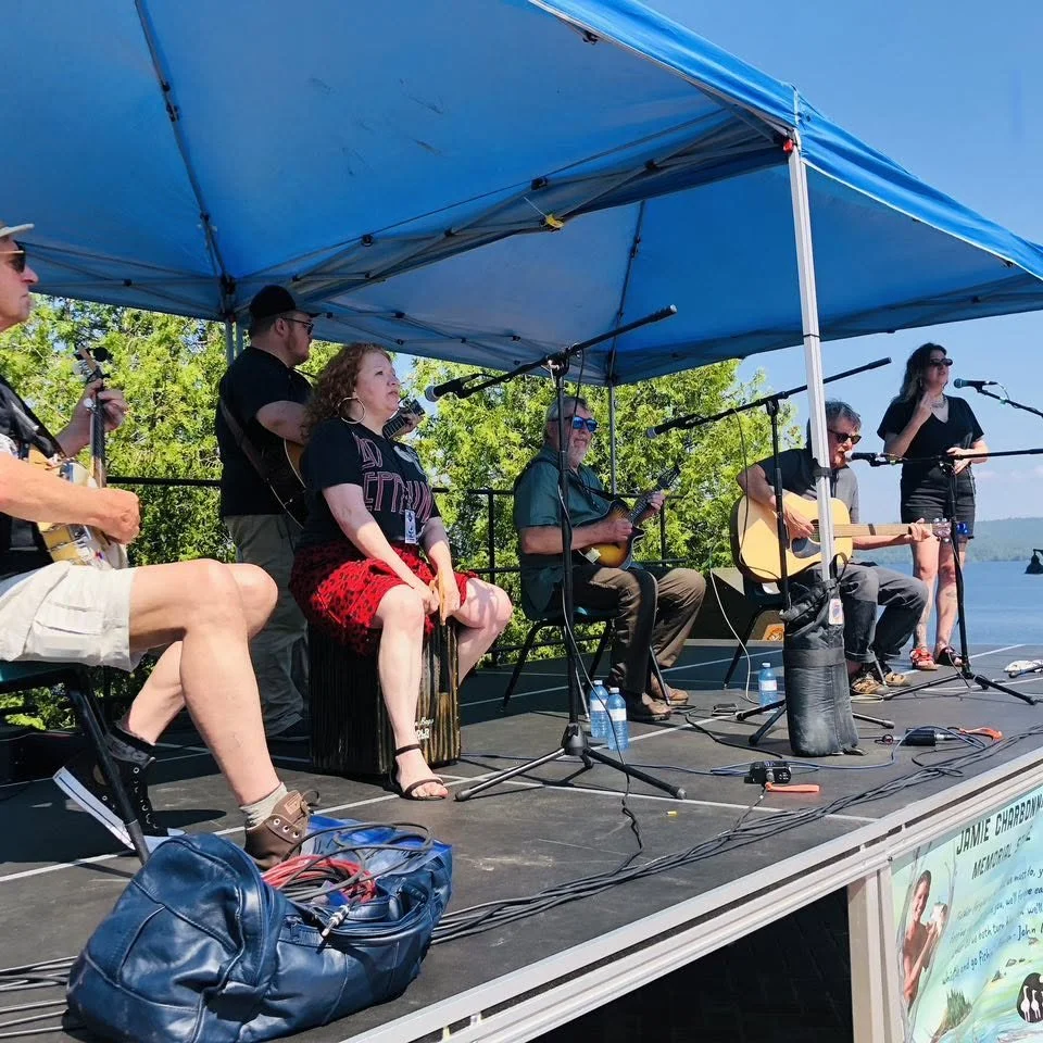 A group of musicians and a singer performing on an outdoor stage under a blue canopy on a sunny day, with trees and water in the background.