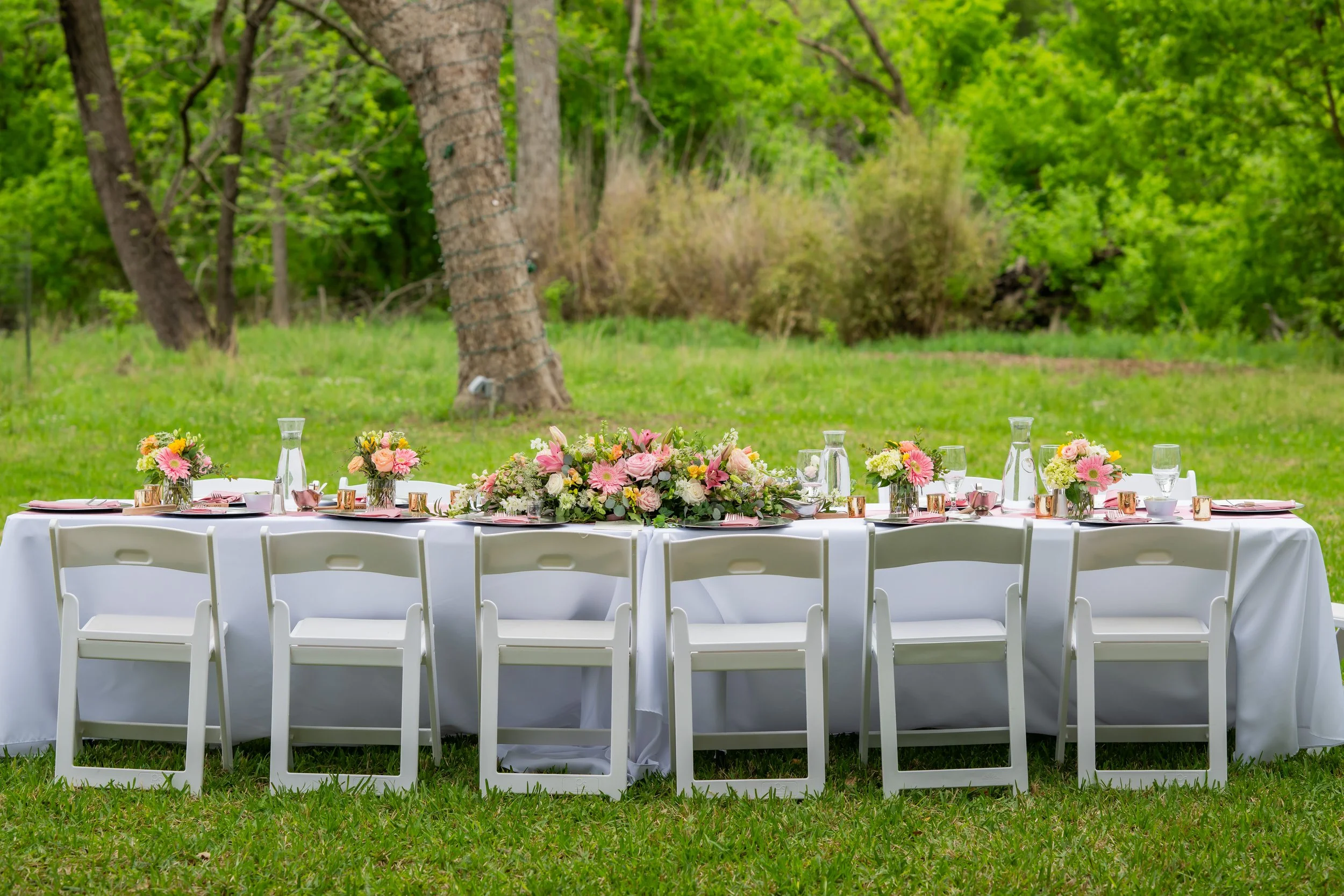 Long outdoor table with white tablecloth, decorated with pink, yellow, and white flowers, set with glasses, plates, and napkins, located in a green grassy area with trees in the background.