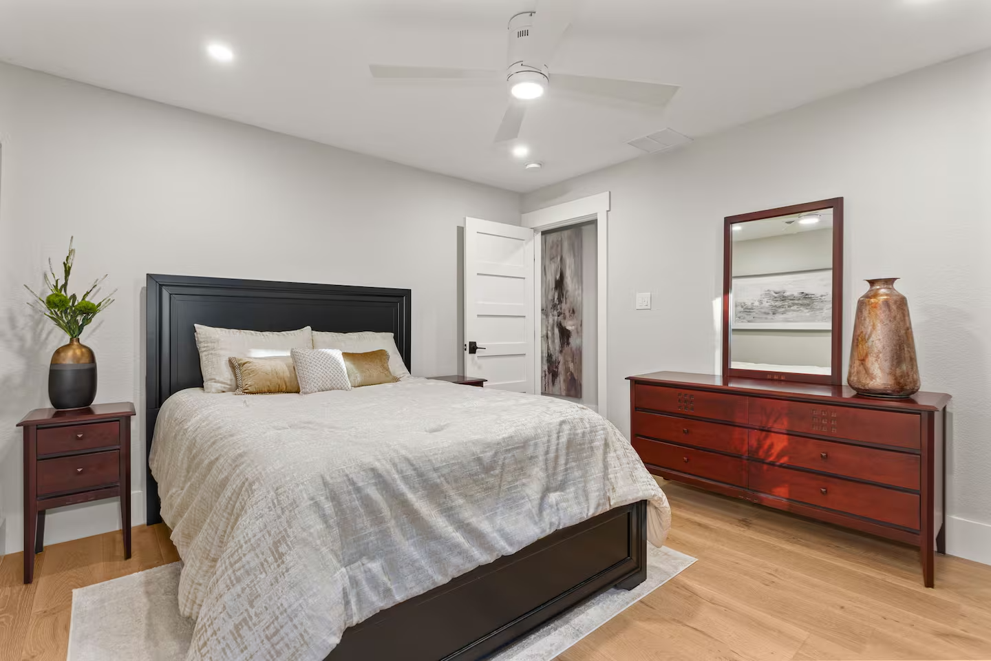 A bedroom with a black bed frame, beige bedding, wooden nightstands, a dresser with a mirror, large vase, and decorative pillows, with a white door and ceiling fan.