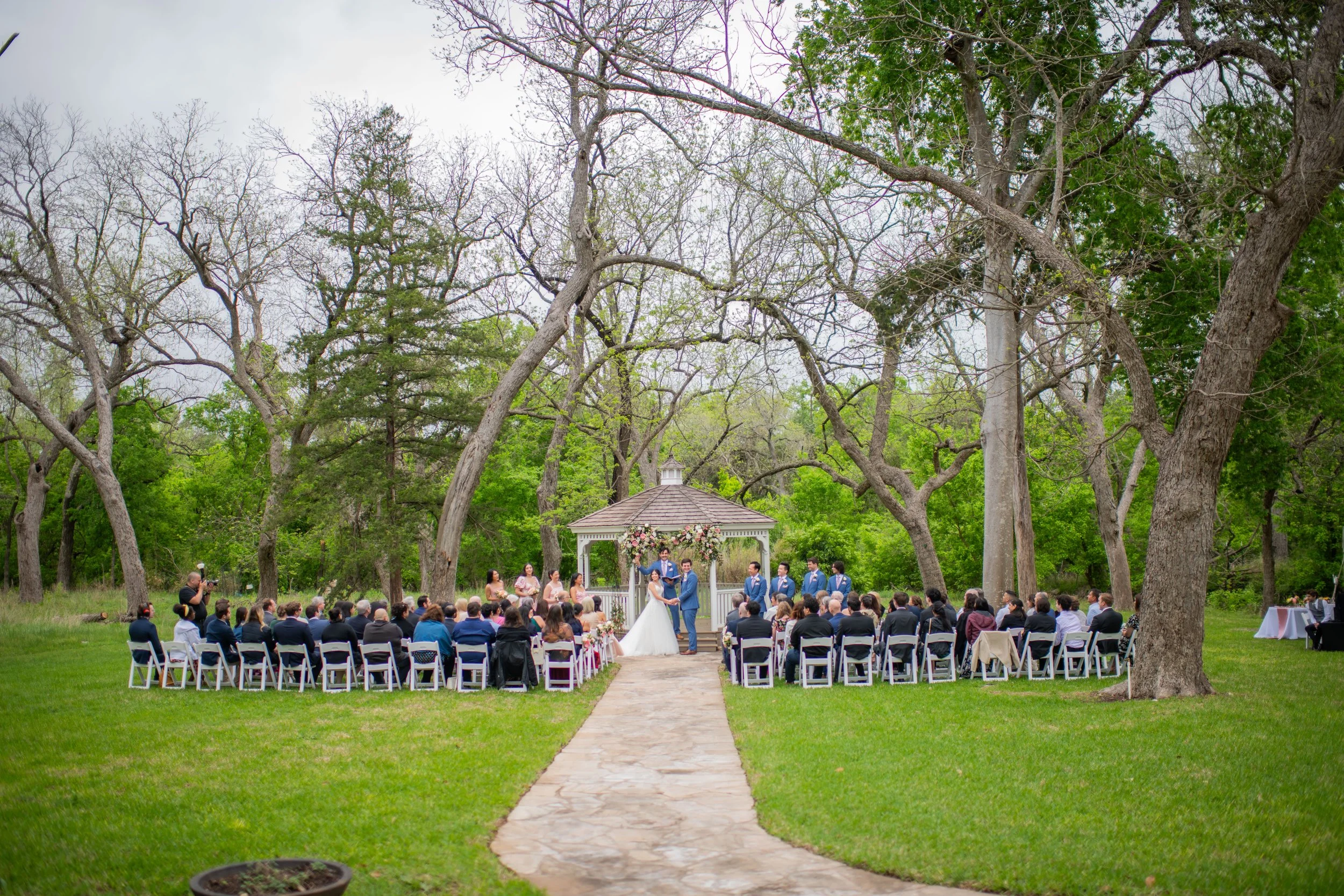 An outdoor wedding ceremony with a bride and groom standing under a gazebo decorated with flowers, surrounded by seated guests. The setting is a park with tall trees and a cloudy sky.