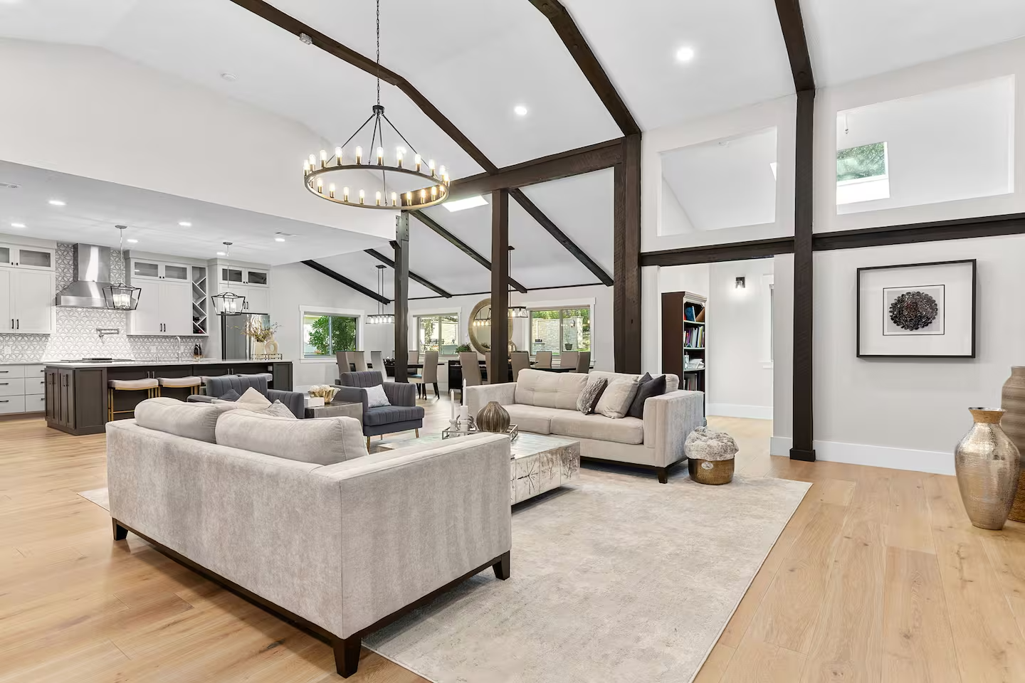Open-concept living room with white sofas, wooden floors, and a kitchen and dining area in the background, decorated with modern lighting and artwork.