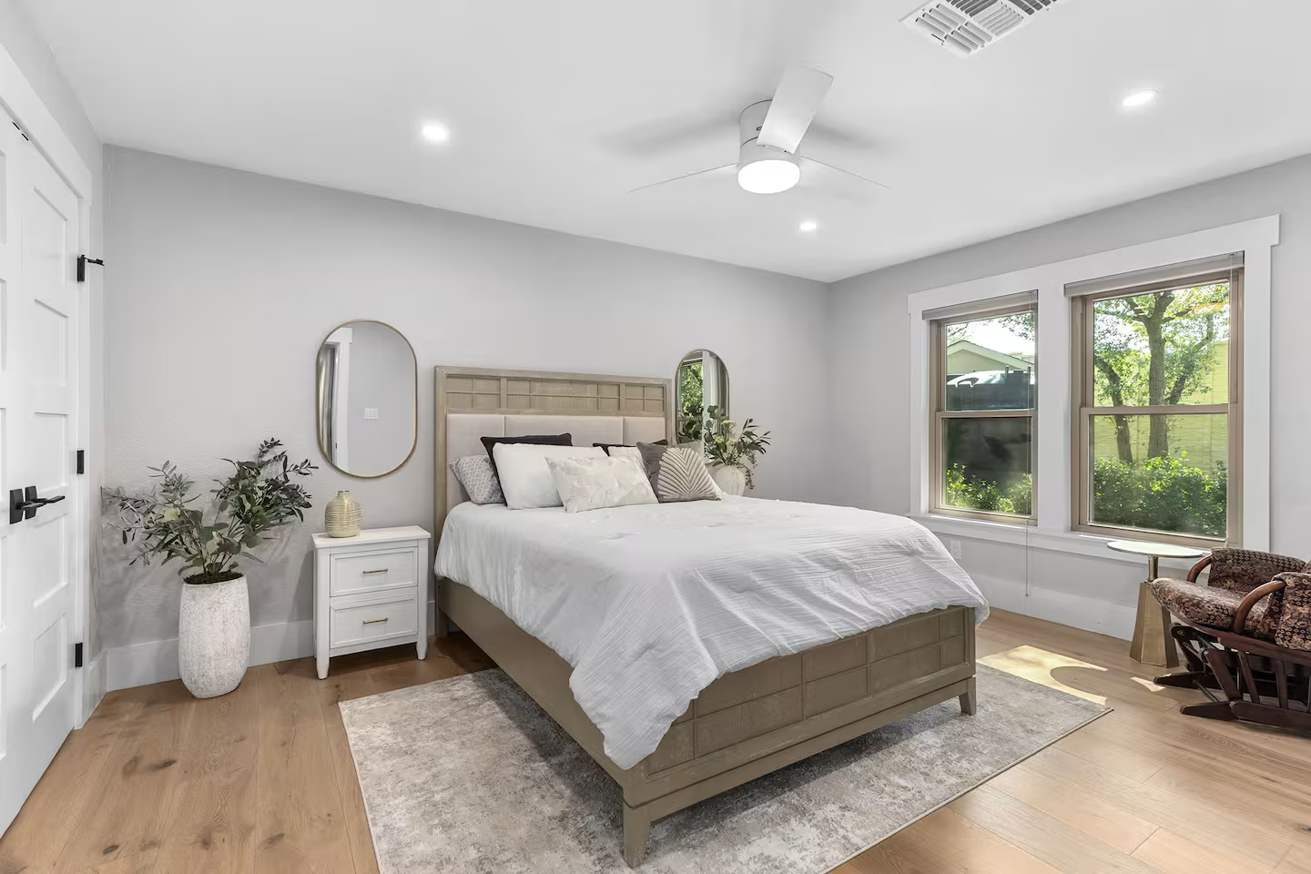 Bright, minimalist bedroom with a large bed, white bedding, wooden furniture, a ceiling fan, large windows with greenery outside, a potted plant, a small white bedside table, and a vintage armchair.