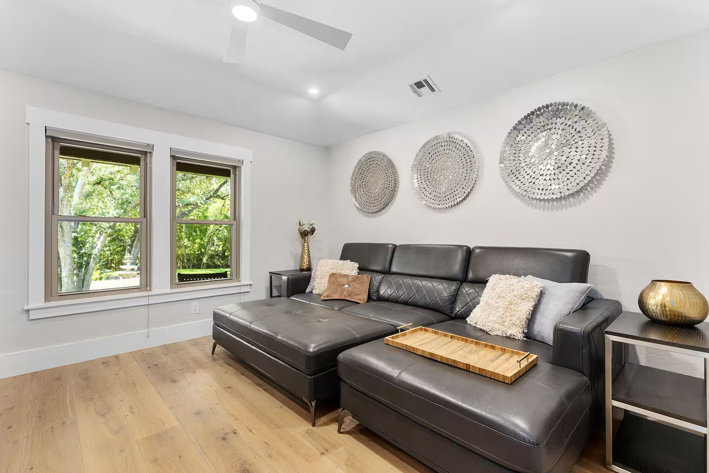 Living room with large black leather sectional sofa, wooden tray on the sofa, decorative pillows, three circular textured wall art pieces, two windows with greenery outside, wooden floor, side tables with vases, ceiling fan, and recessed lighting.
