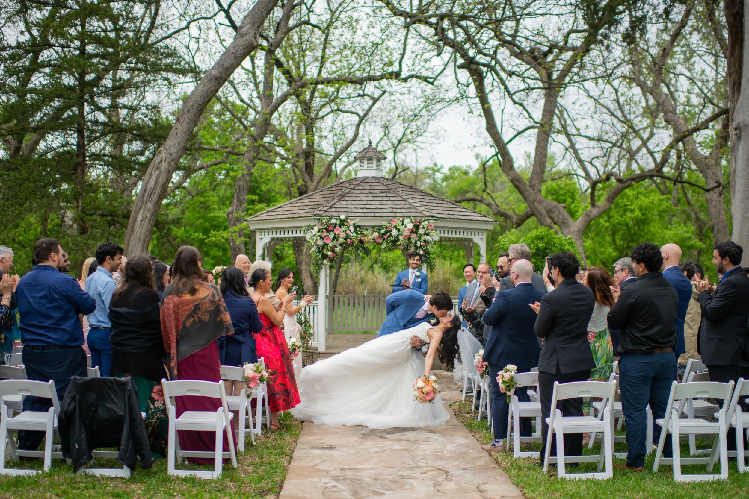 A wedding ceremony outdoors with a female bride in a white gown and a male groom in a blue suit, surrounded by guests. They are under a gazebo decorated with pink and white flowers, in a park with tall trees and green foliage.