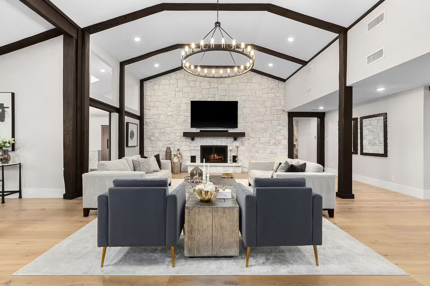 Modern living room with vaulted ceiling, wood beams, stone fireplace, flat screen TV, white and gray furniture, gold decor accents, and a large chandelier.