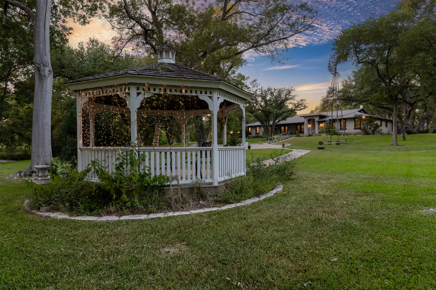 A white gazebo decorated with string lights situated on a grassy lawn, with a winding path leading to a modern house in the background during sunset.