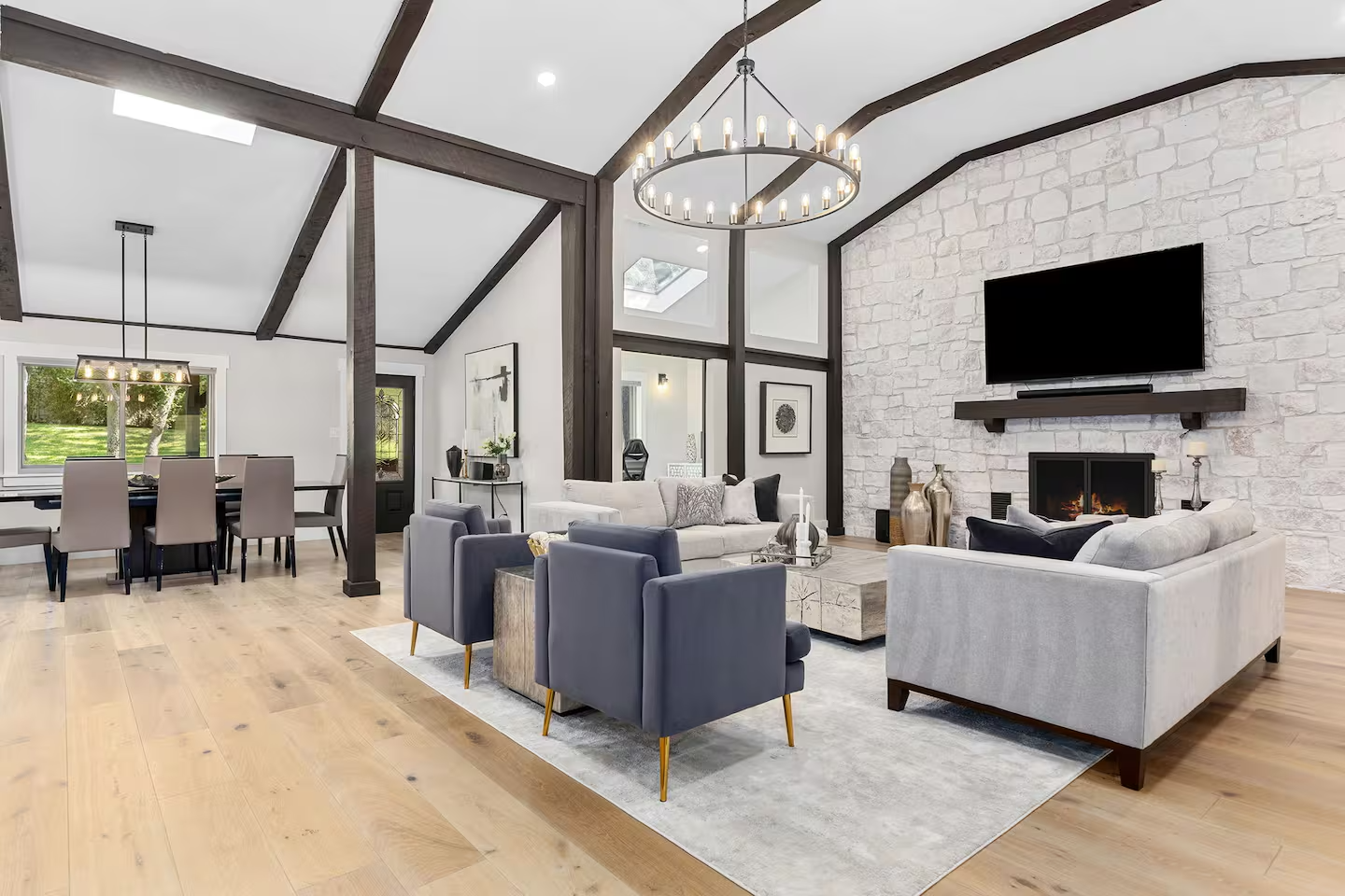 Living room with white stone fireplace, flat-screen TV, modern furniture including beige and dark blue armchairs and beige sofas, wooden beams on ceiling, and hardwood flooring.