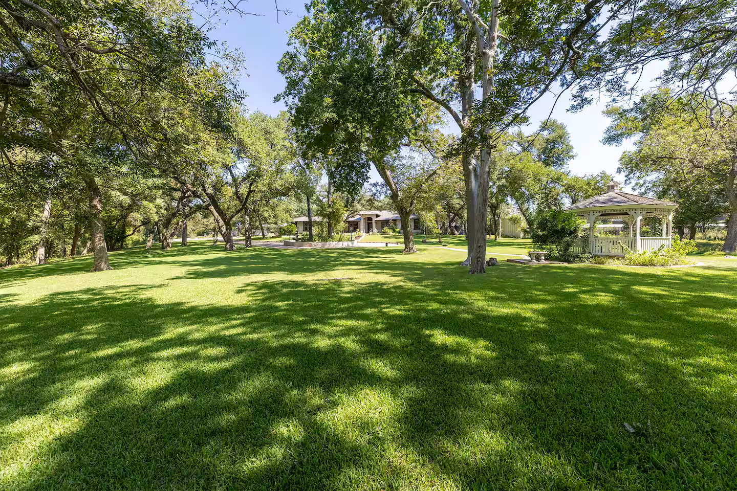 Wide view of a lush green park with tall trees, a white gazebo, and a house in the background, under a clear blue sky.