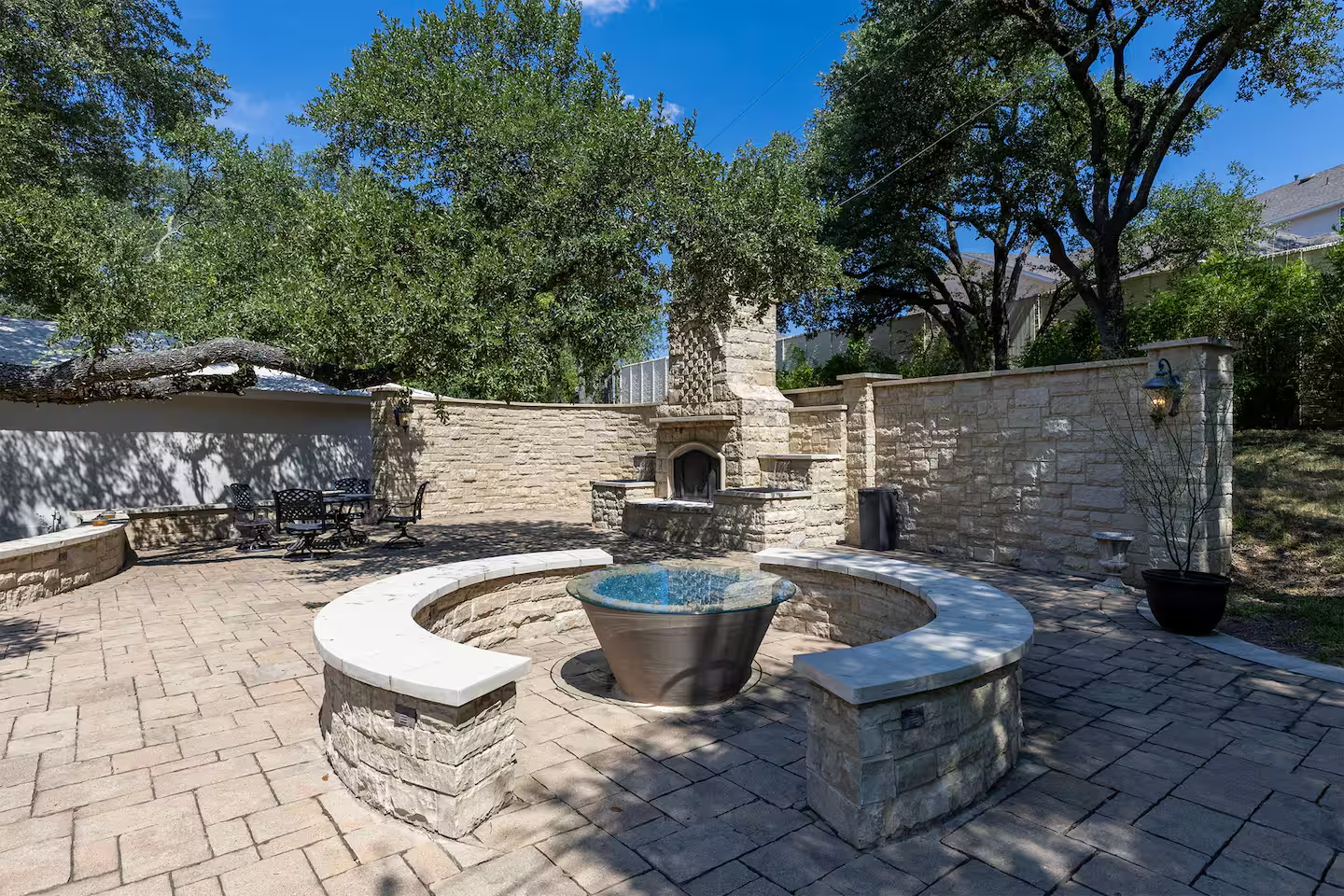 Appears to be a backyard patio with brick pavers, a round fire pit with a concrete bench surrounding it, trees providing shade, and a stone fireplace on the back wall. There are outdoor chairs and a table on the left side, with a large potted plant and a wall-mounted lantern on the right.