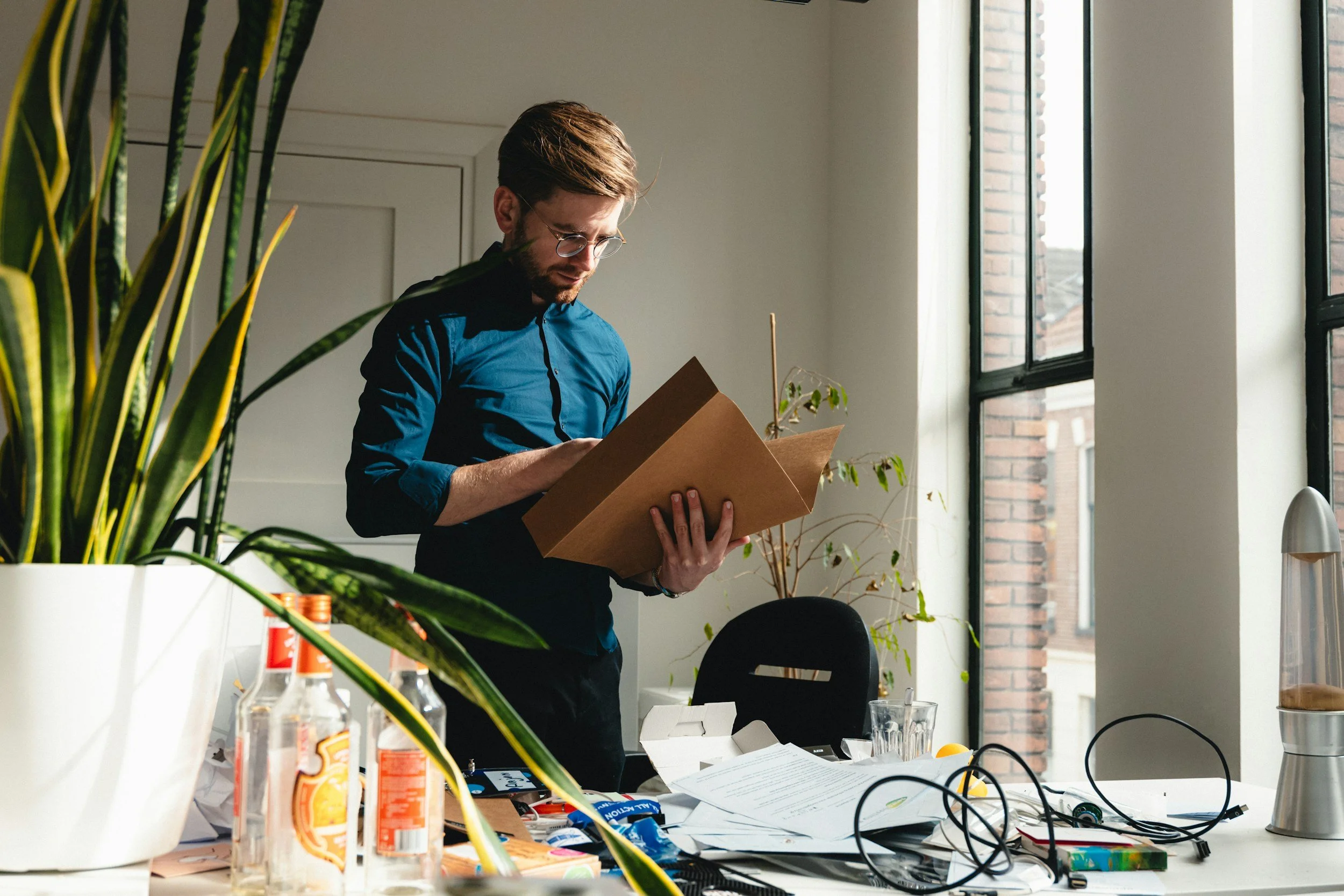 A young man with glasses and brown hair, wearing a blue shirt, is standing at a cluttered table examining a cardboard box. The table is filled with papers, electronic devices, and bottles. Bright natural light enters the room through large windows, and there are potted plants nearby.
