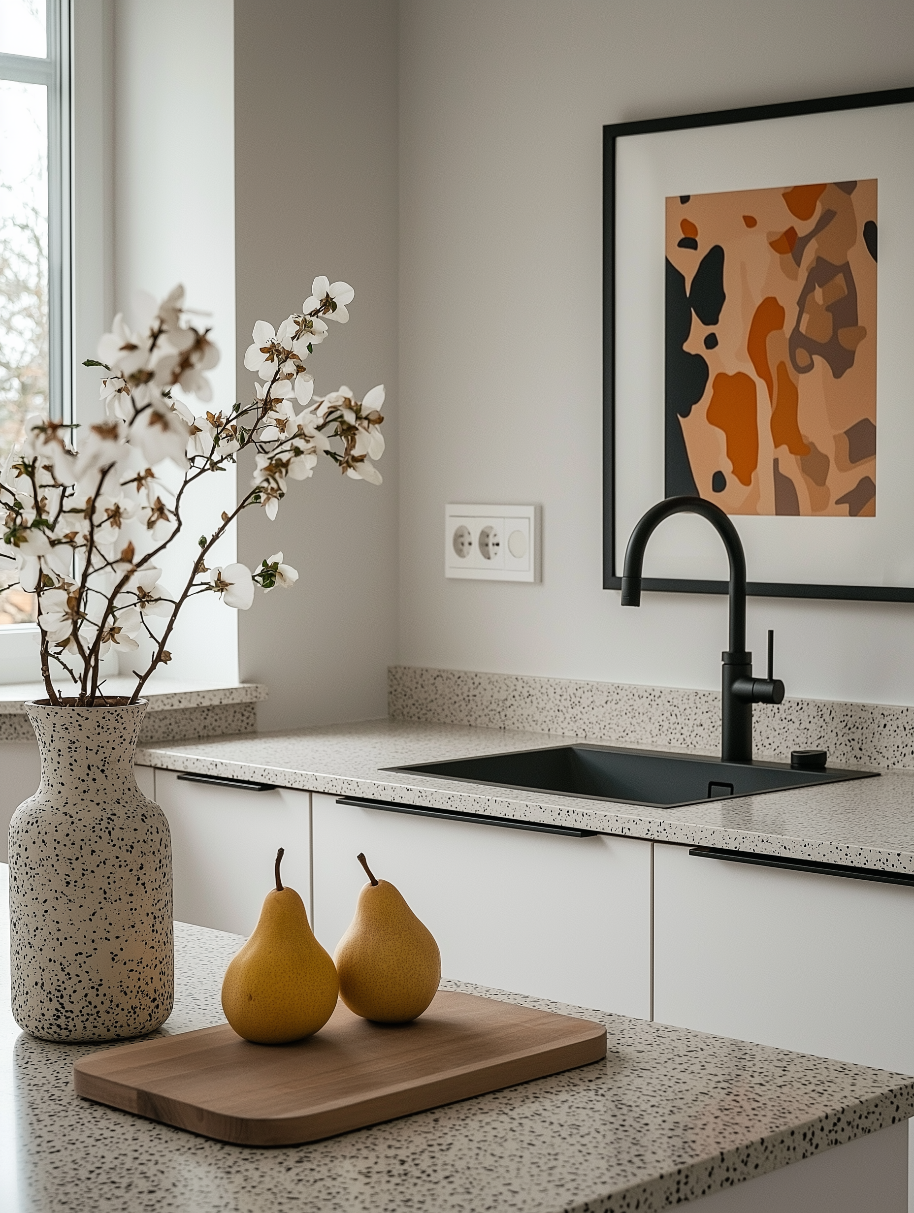 A modern small kitchen featuring speckled countertops, a matte black sink with matching faucet, and a wooden cutting board with two pears. The space is styled with a speckled ceramic vase holding white flowers and abstract wall art in neutral tones.