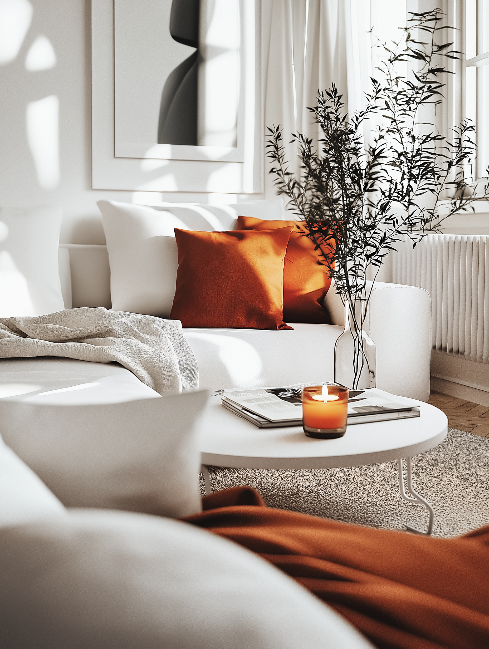 Bright and cozy living room with vibrant rust colored pillows, a white sofa, minimalist decor, a glowing candle, and a modern table adorned with greenery for a warm and inviting atmosphere.