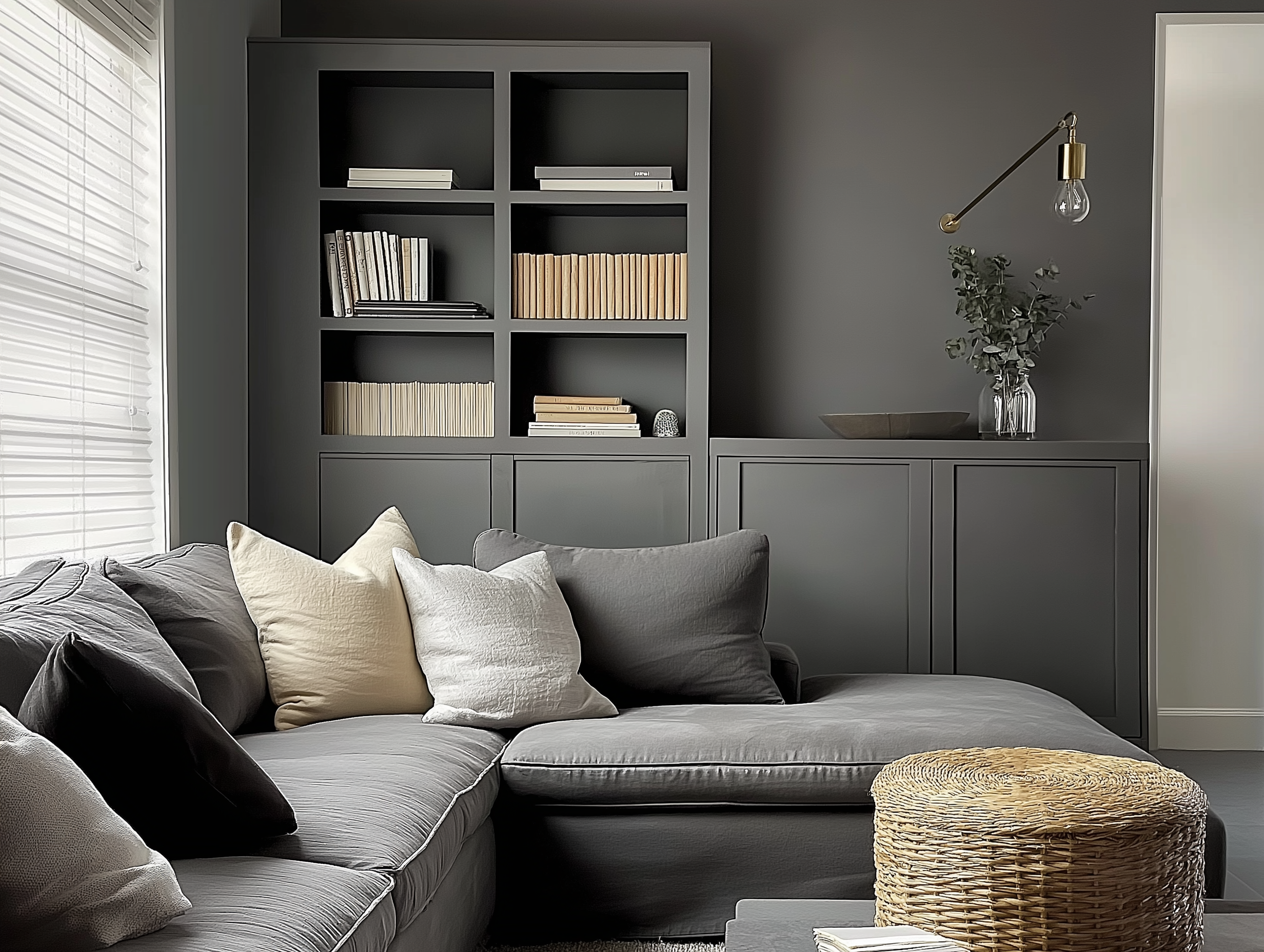 A chic gray living room featuring Farrow & Ball’s Down Pipe painted built-in shelves, a cozy gray sectional, neutral throw pillows, a woven ottoman, and a brass wall sconce for an elegant yet modern aesthetic.