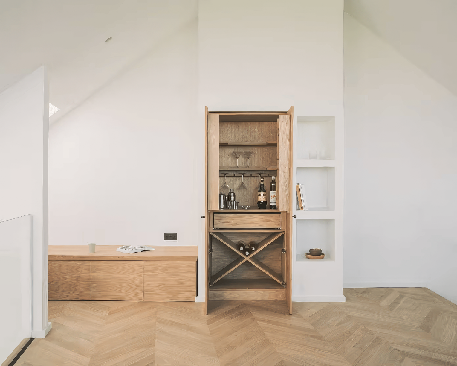 Minimalist built-in wooden bar cabinet with open doors revealing wine bottles, glasses, and cocktail accessories, set against a clean white wall and herringbone parquet flooring.