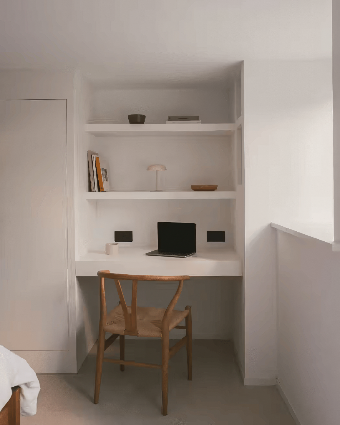 Small minimalist desk setup with floating shelves, white built-in desk, laptop, and natural wishbone chair under a window.