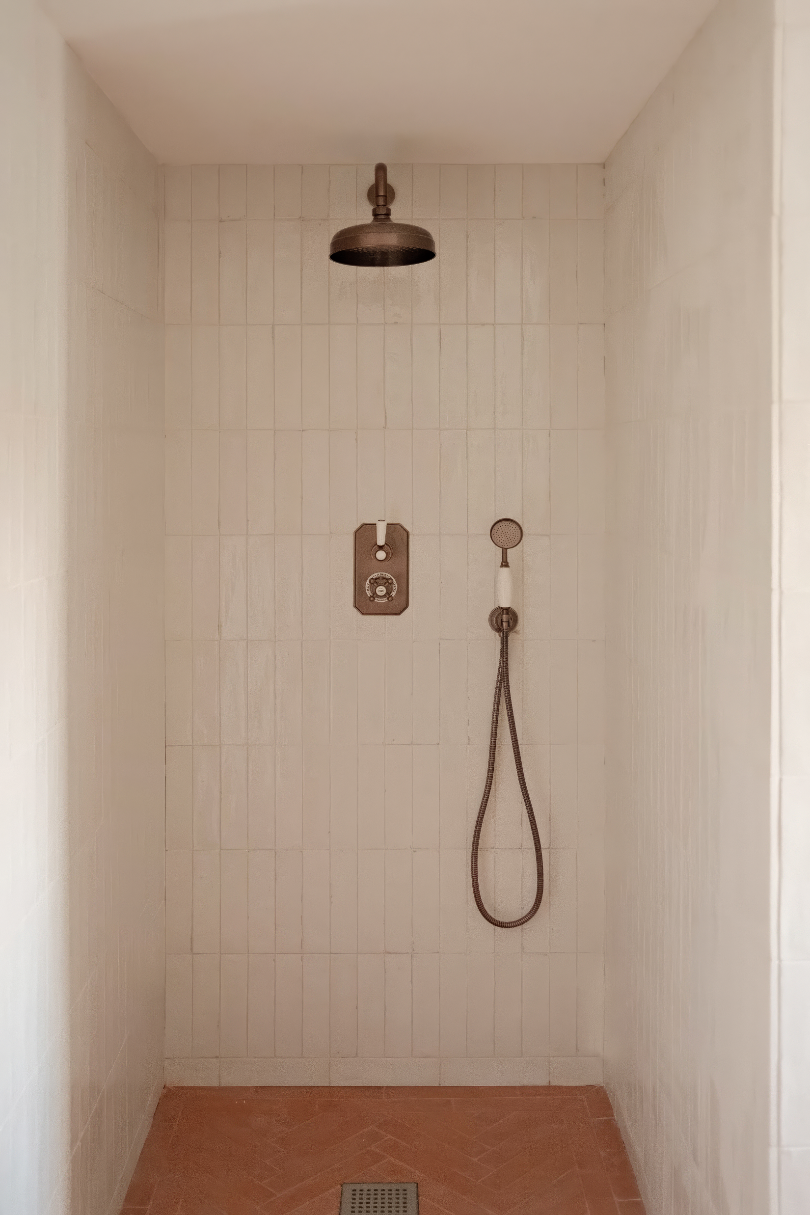 Walk-in shower with vertically stacked cream tiles, vintage bronze rainfall showerhead, and terracotta herringbone floor.