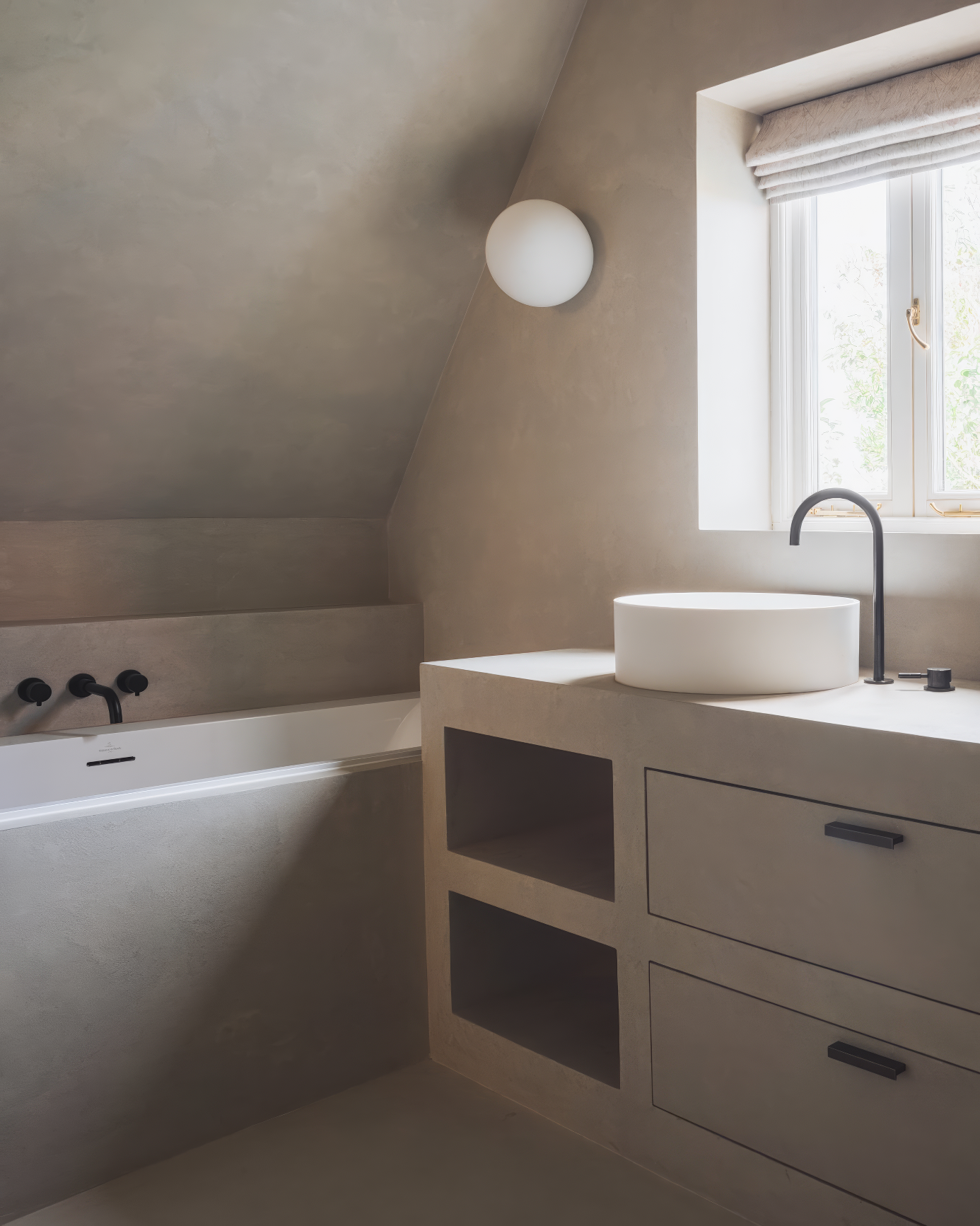 Minimalist bathroom with soft taupe-toned plaster walls, a white freestanding bathtub, and a round vessel sink beneath a small window.