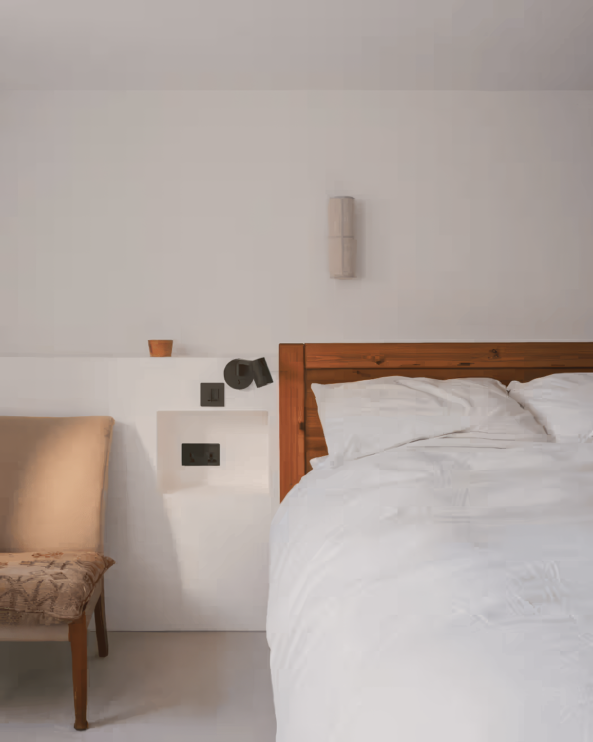 Cozy bedroom corner with white walls, vintage armchair, wood-framed bed, and inset shelf with black and terracotta accents.