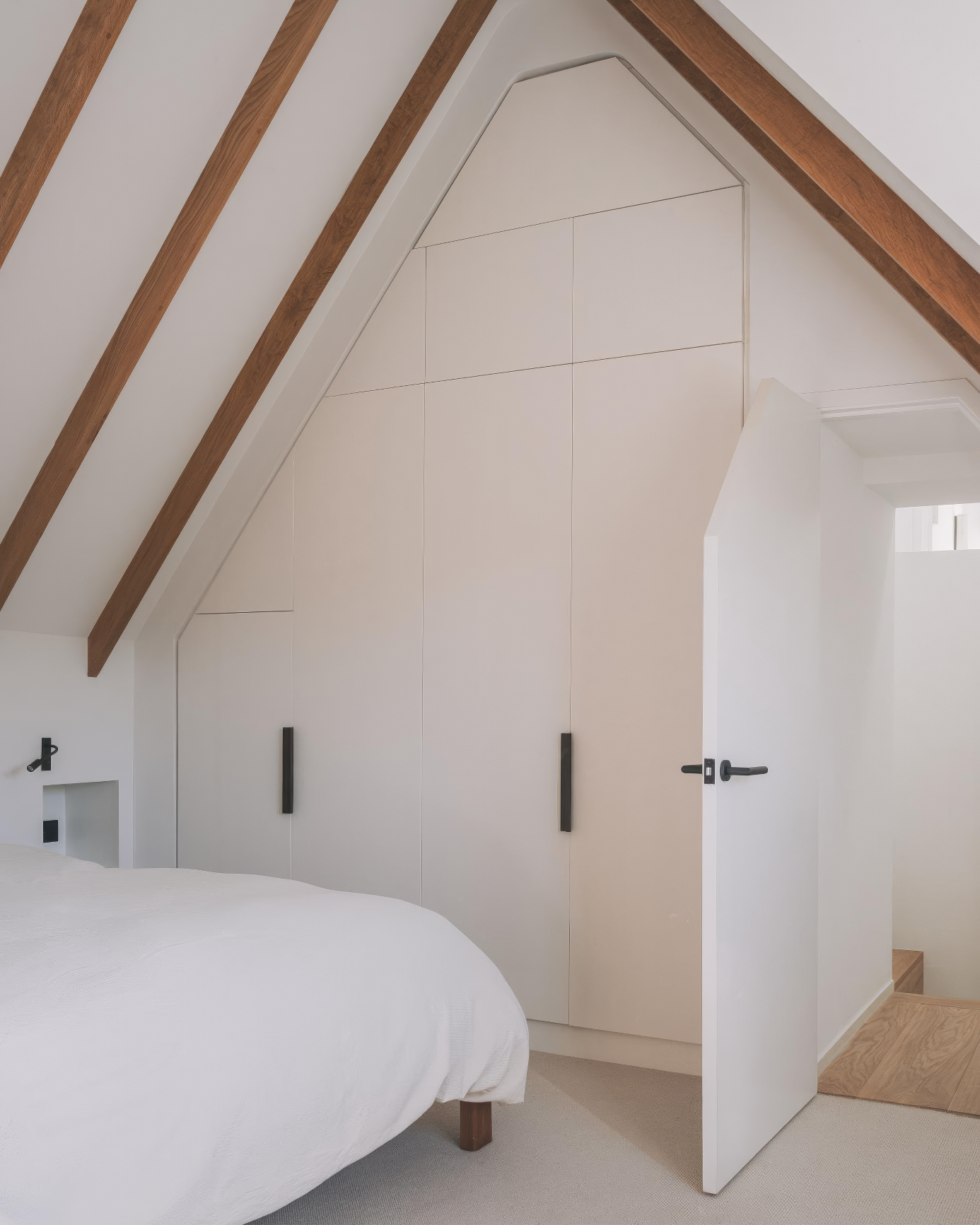 Custom-built wardrobe fitted into a gabled ceiling alcove with sleek beige doors and contrasting black handles, located in a modern loft bedroom with exposed wood beams.