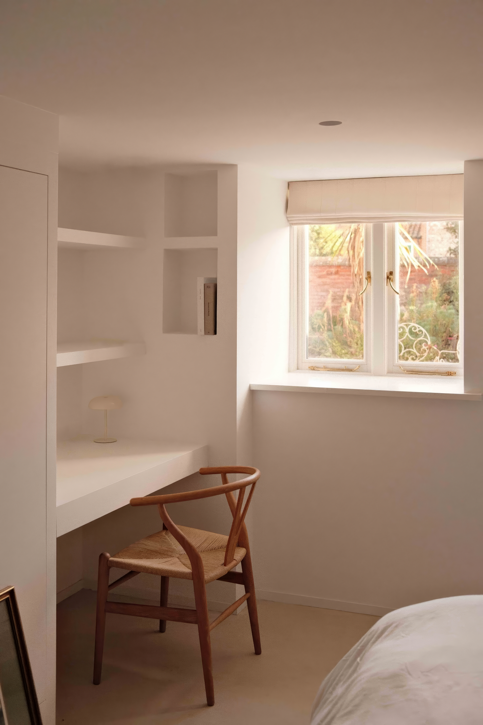 Small home workspace with built-in white desk, bookshelves, and a natural wood wishbone chair, lit by a window with Roman shade.