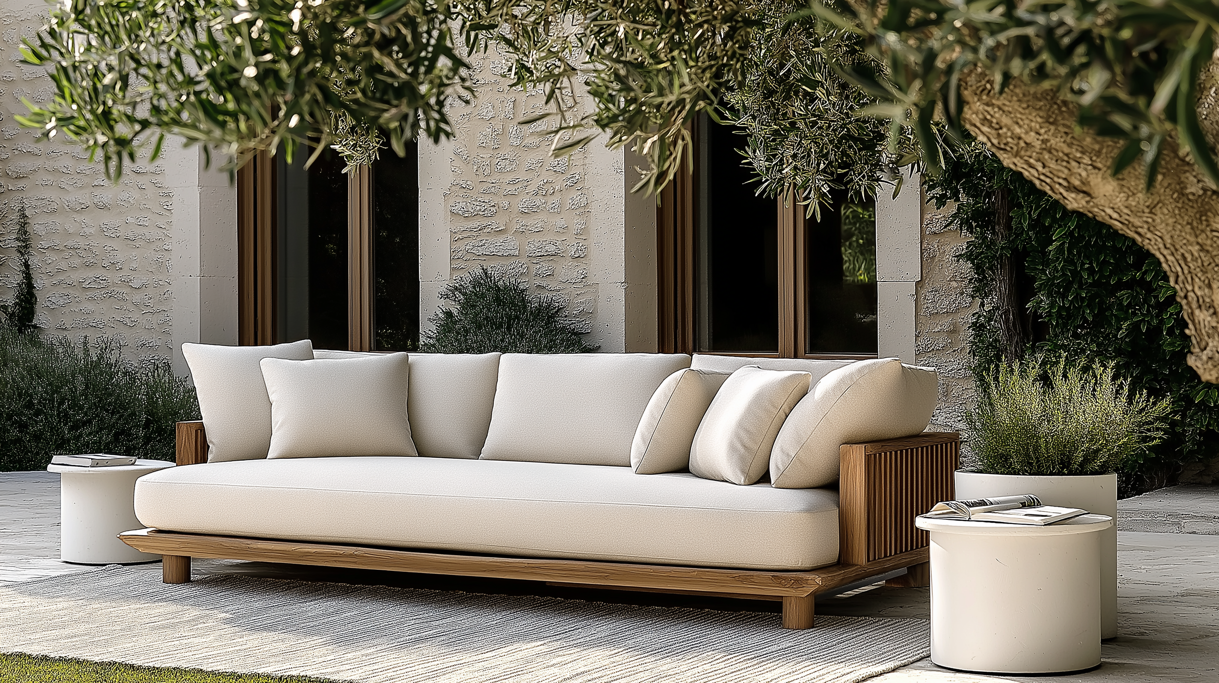 A modern outdoor sofa with a wooden frame and beige cushions, placed on an area rug in neutral tones, surrounded by white concrete side tables with green plants, under the shade of olive trees.