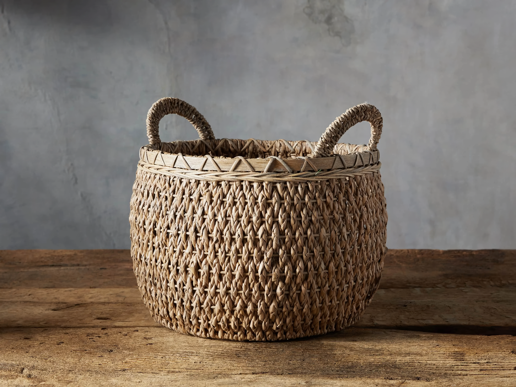 Round Entry Basket handwoven from natural seagrass kalas. Features a subtle natural or dark finish set on a rustic wooden floor with a grey background.