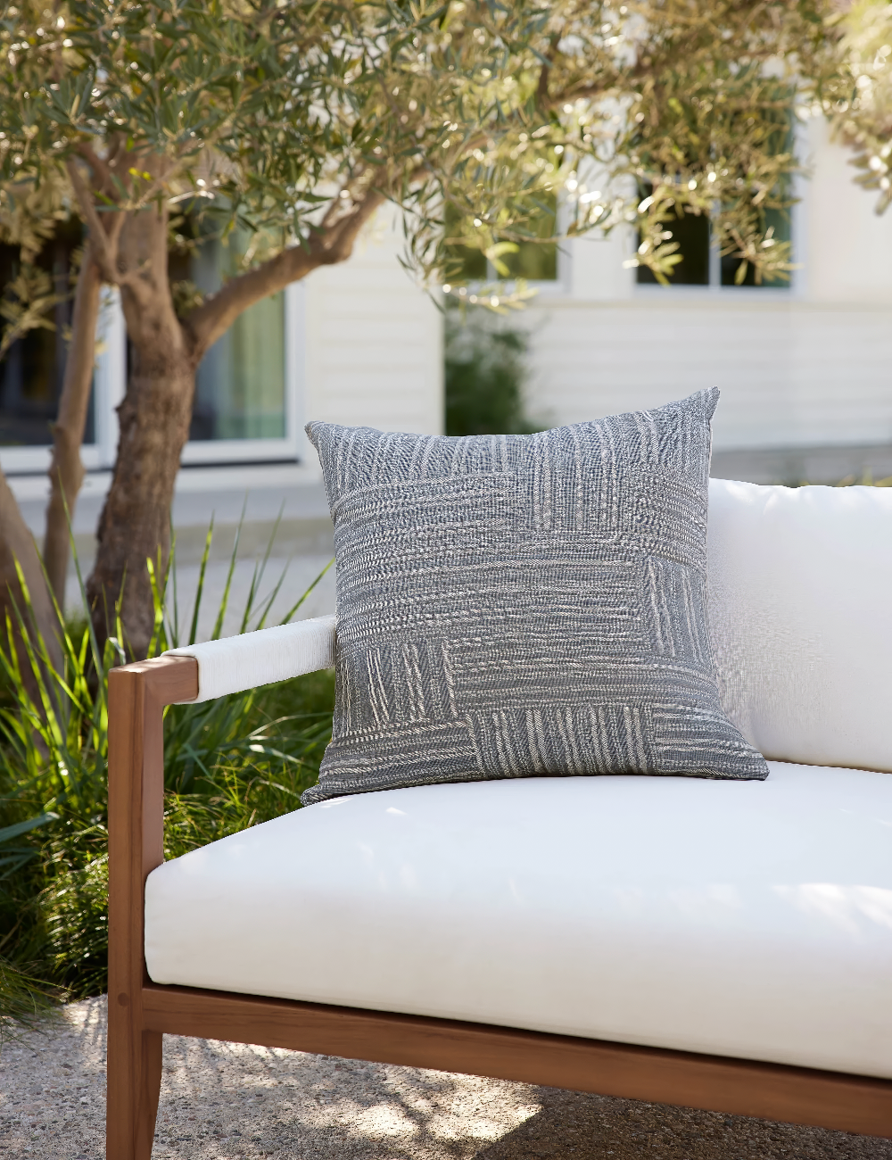 Abstract grey and cream crosshatch pattern of this throw pillow is inspired by traditional kuba cloth. The throw pillow is in a garden setting on an outdoor sofa.
