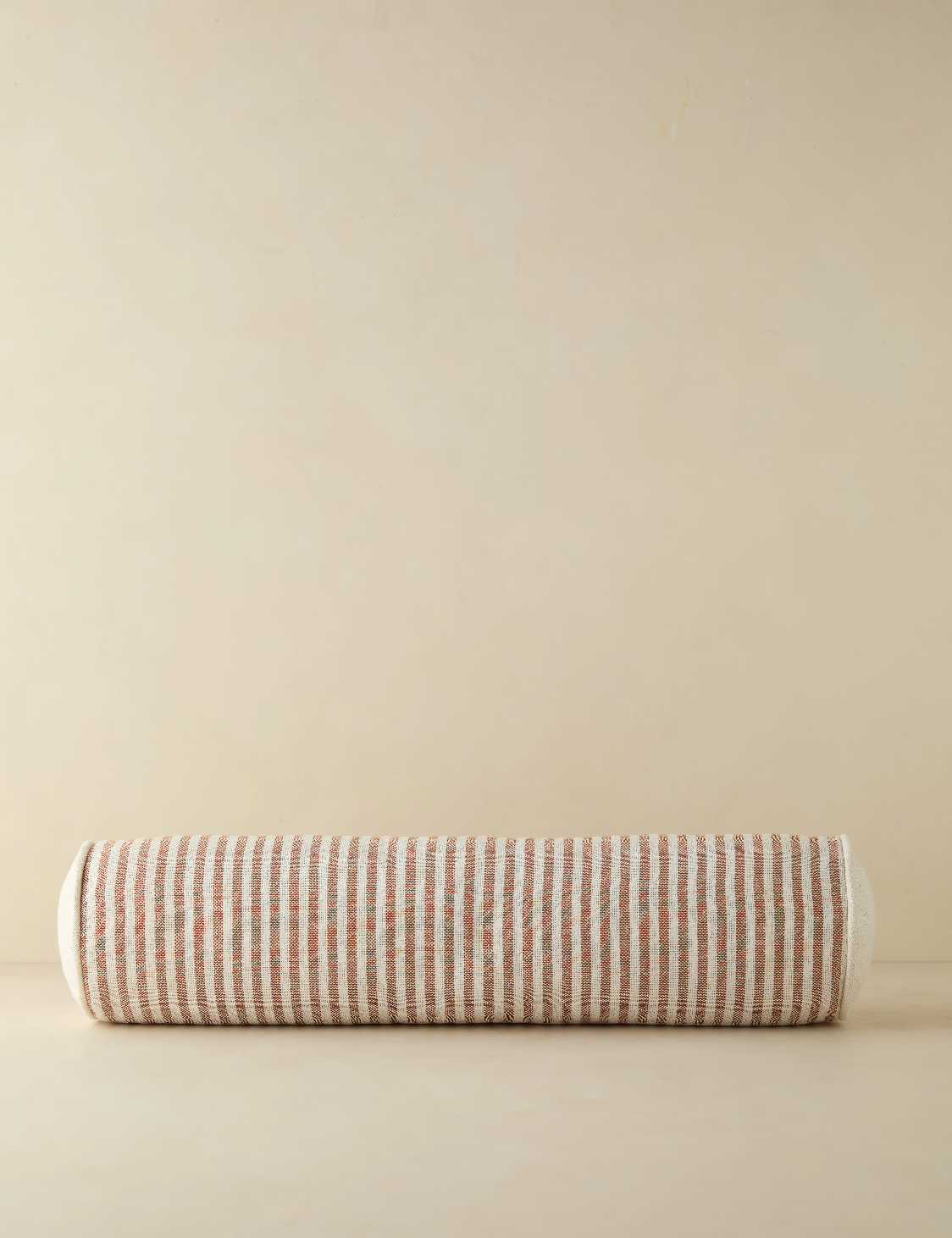 Neutral-toned striped bolster pillow with a cylindrical shape, featuring alternating soft beige and white stripes. The pillow rests against a light beige background, exuding a minimal and cozy aesthetic.