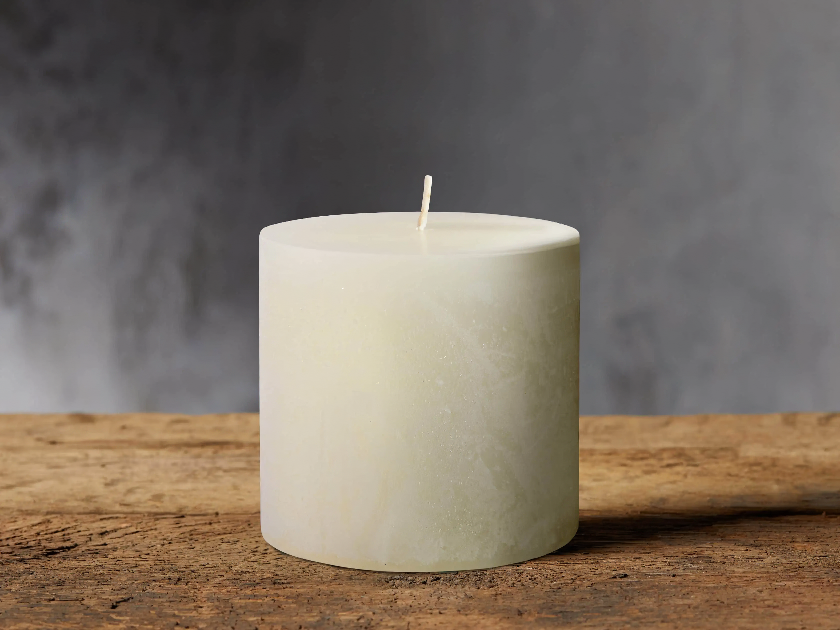 Simple off-white pillar candle with a smooth, slightly marbled texture, placed on a rustic wooden surface against a moody gray background. The minimalist design adds a warm, calming ambiance to any space.