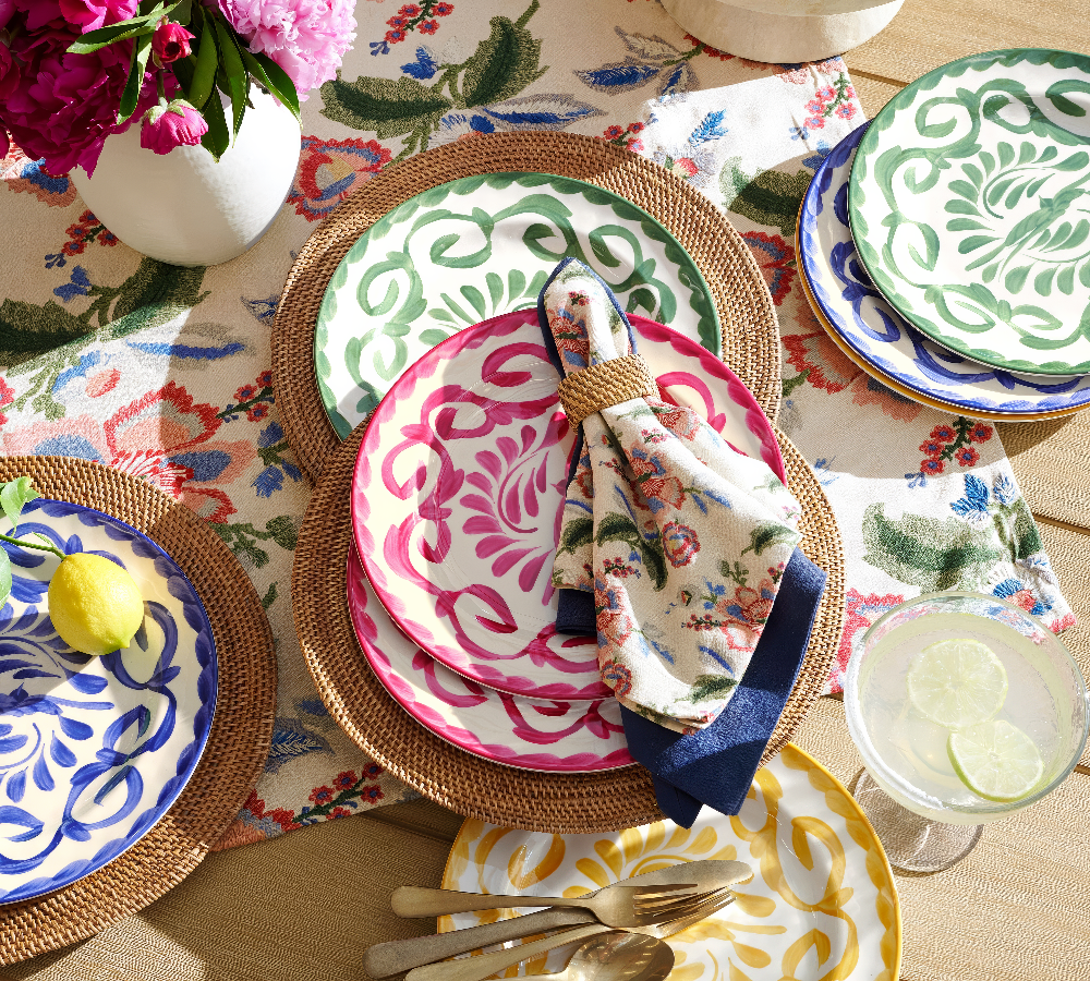 Vibrant outdoor dining setup featuring colorful melamine dinner plates with intricate swirling patterns in pink, green, blue, and yellow. The plates are arranged on woven rattan placemats atop a floral tablecloth, accompanied by a decorative napkin.