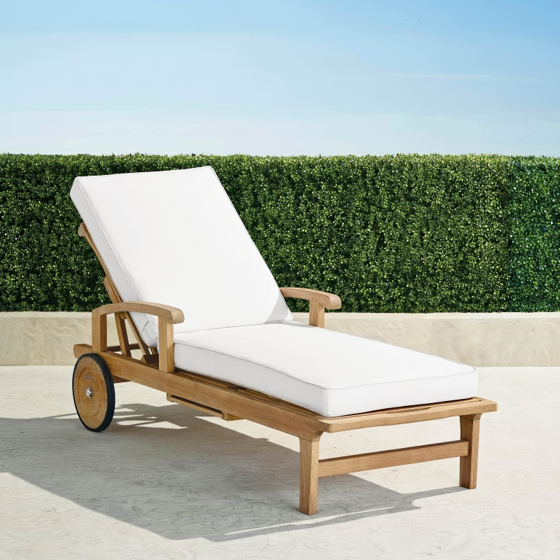 Outdoor teak chaise lounge with plush white cushions, featuring an adjustable backrest and smooth curved armrests. The lounge is set on a sunlit patio with a neatly trimmed green hedge and a clear blue sky in the background.