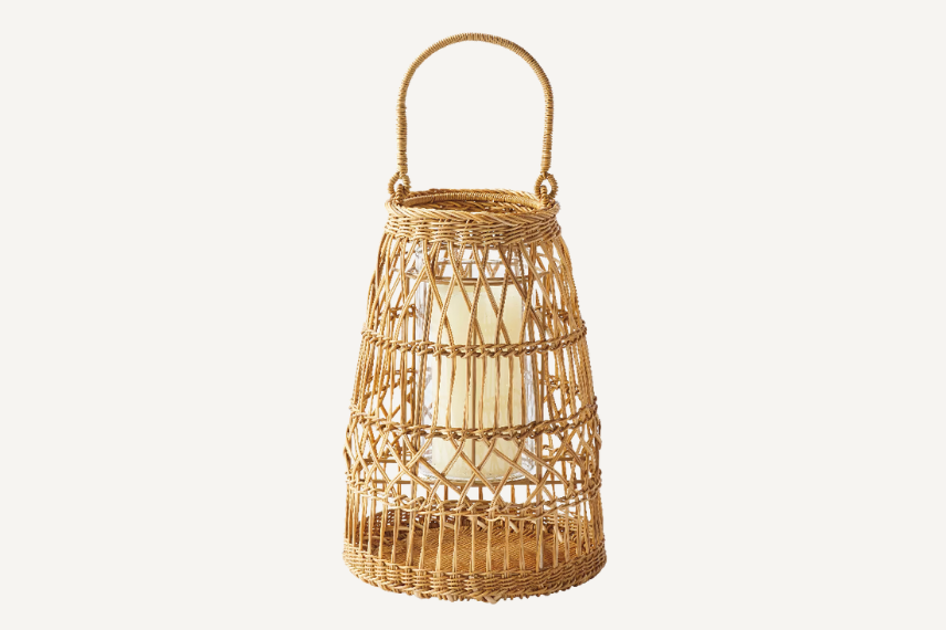 Woven rattan lantern with an intricate open-weave design, featuring a sturdy curved handle and a glass insert holding a candle. The natural texture and warm tones make it a perfect accent for cozy outdoor gatherings or ambient indoor lighting.