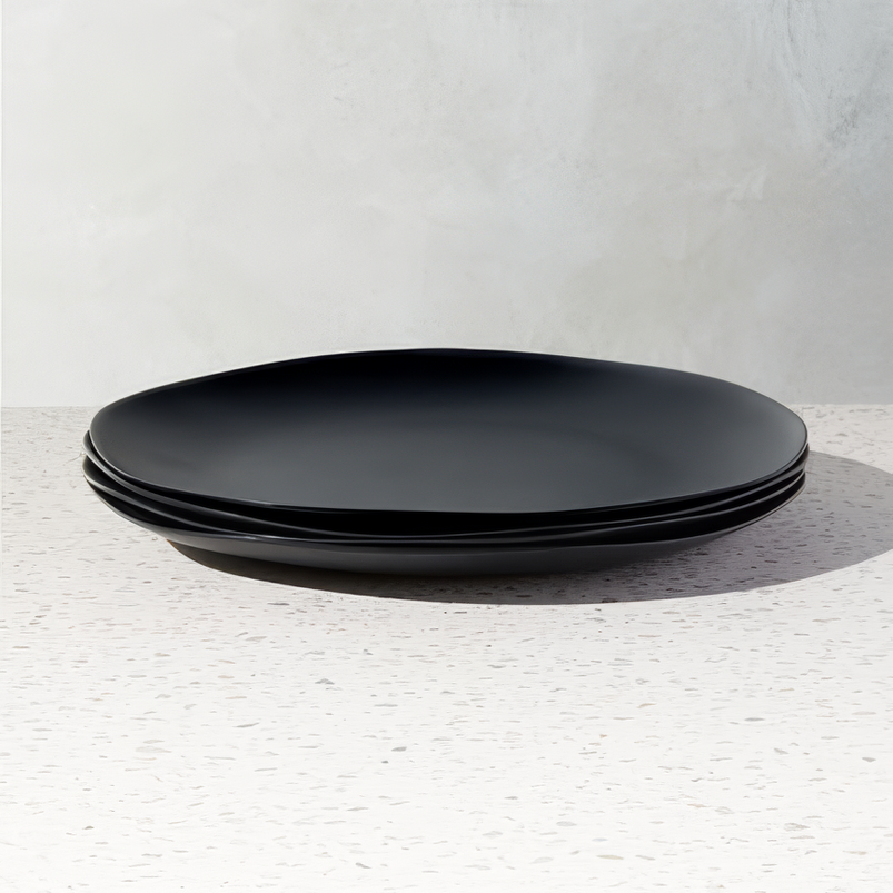 Set of three matte black melamine dinner plates with a modern, organic shape, stacked neatly on a speckled white surface. The sleek, minimalist design adds a contemporary touch to any dining setting.