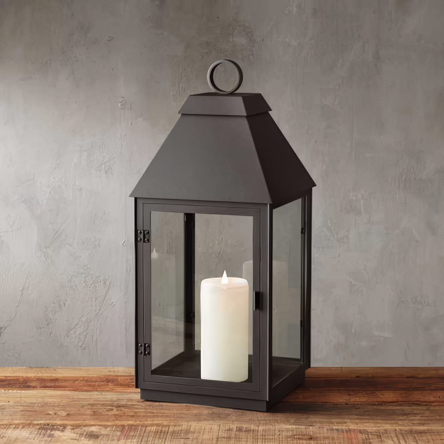 Classic black metal lantern with a peaked roof and glass panels, featuring a white pillar candle inside. The lantern sits on a rustic wooden surface against a textured gray wall, blending modern elegance with a timeless aesthetic.