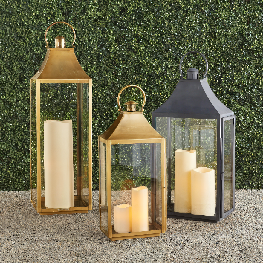 Set of three elegant outdoor lanterns in brass and black metal finishes, each featuring glass panels and glowing pillar candles inside. Placed against a lush green hedge, these lanterns add a warm and sophisticated touch to any outdoor setting.