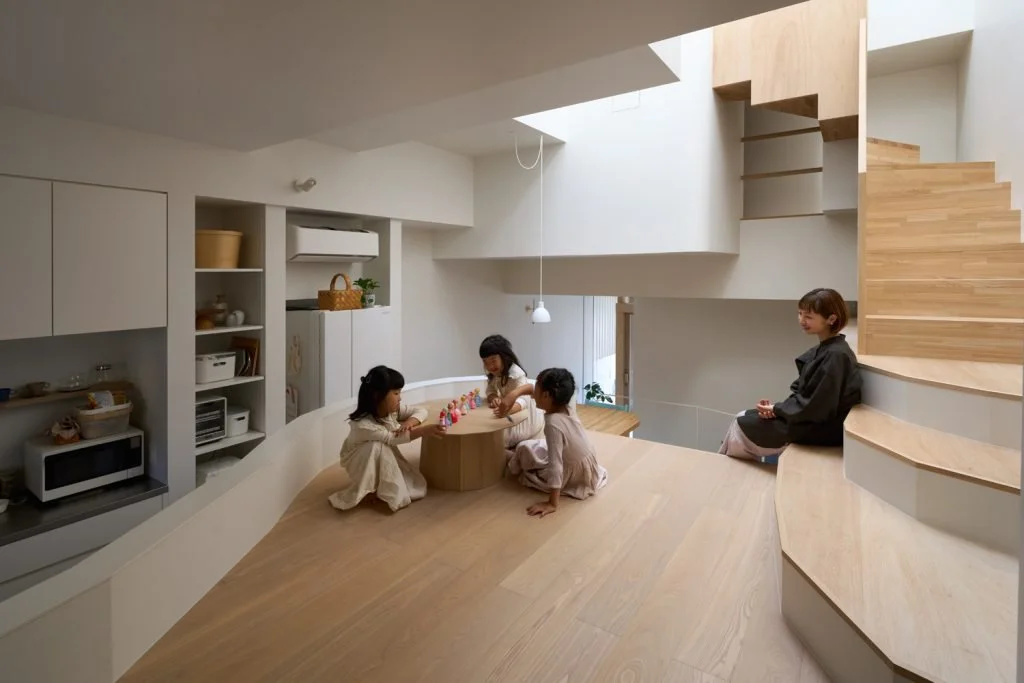 Minimalist Japanese kitchen adjoining a raised living space where children play and a mother sits on built-in bench stairs.