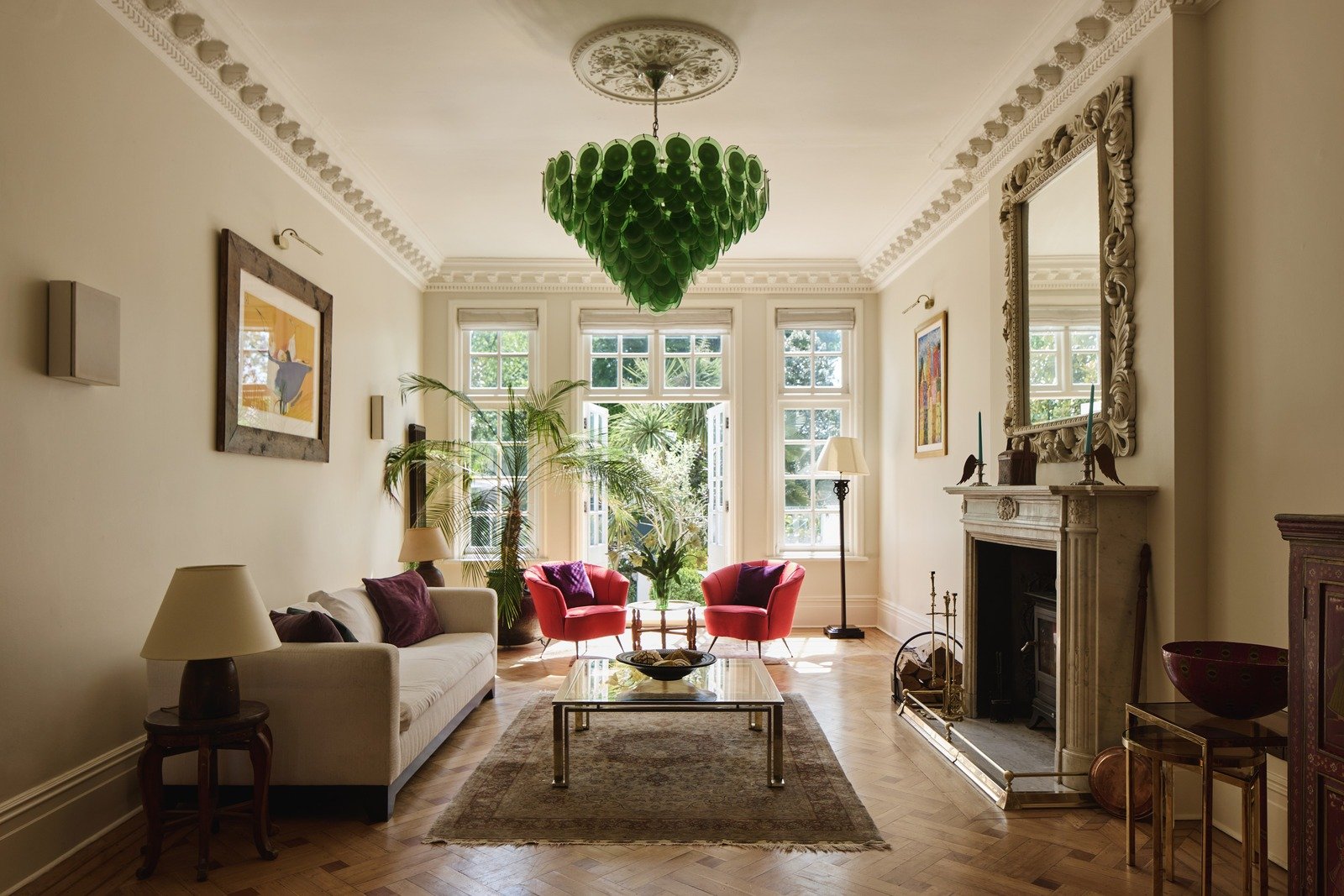Bright and stylish living room with parquet flooring, ornate marble fireplace, modern green glass chandelier, and eclectic furnishings blending classic and contemporary decor.