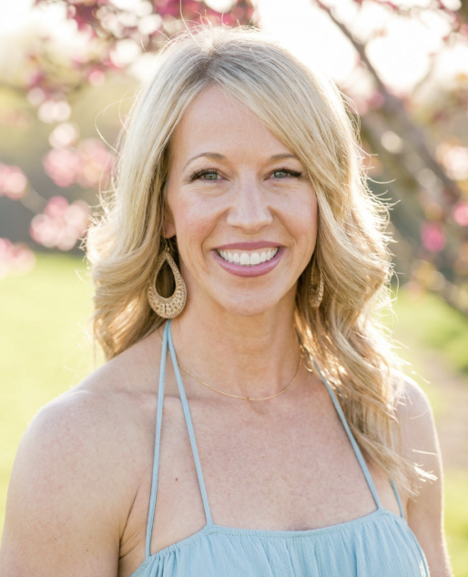 A smiling woman with long blonde hair wearing a light blue dress and large earrings outdoors, with pink flowering trees in the background.
